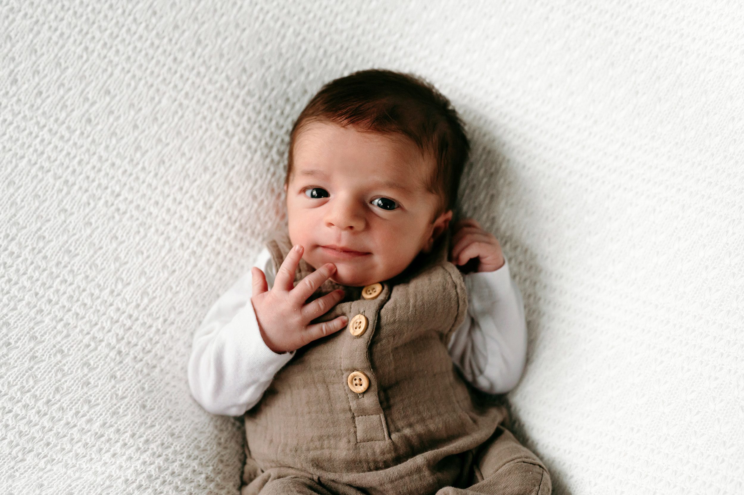 a baby boy wearing a long sleeve onesie and white overalls laying on a textured white backdrop and touching his chin as he gazes up at the camera during a newborn photos session