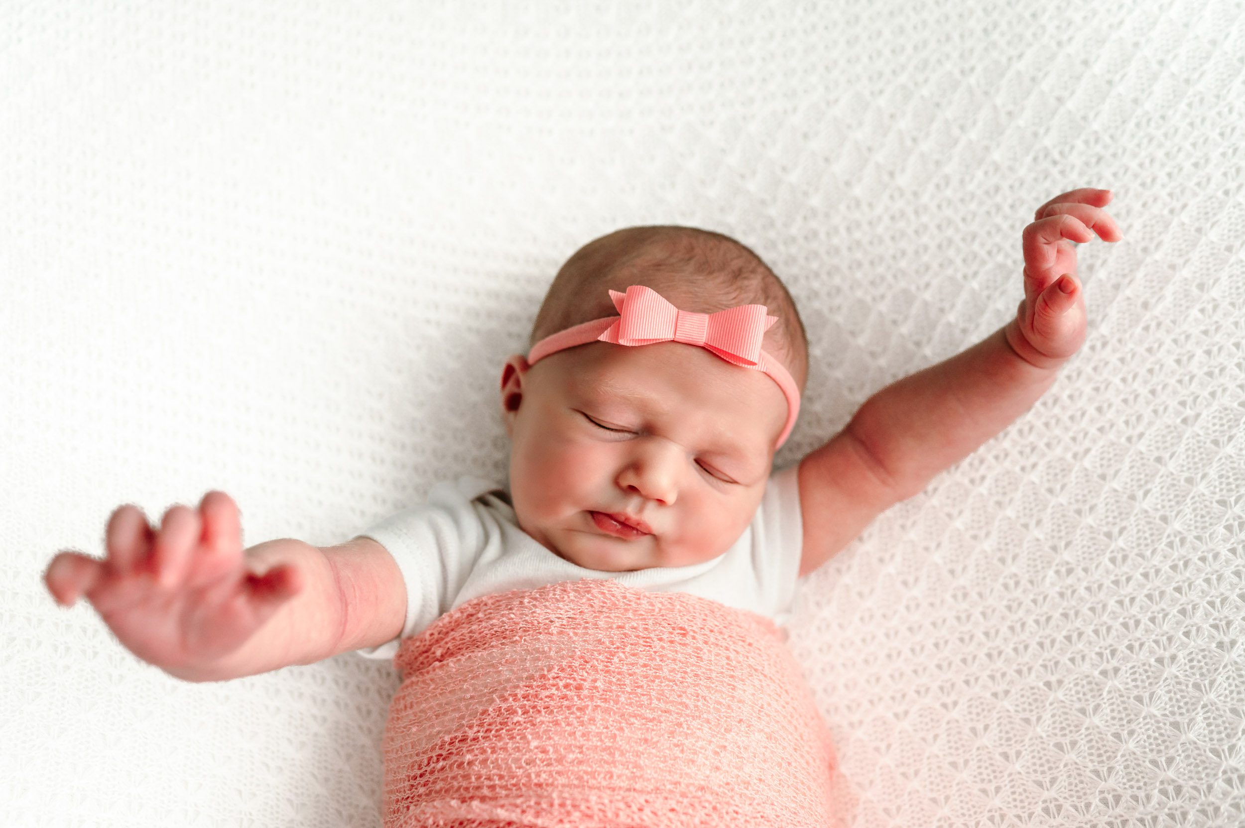 a baby girl wearing a white onesie and pink headband wrapped in a matching pink swaddle stretching her arms out wide as she wakes up from a nap during a newborn photos session