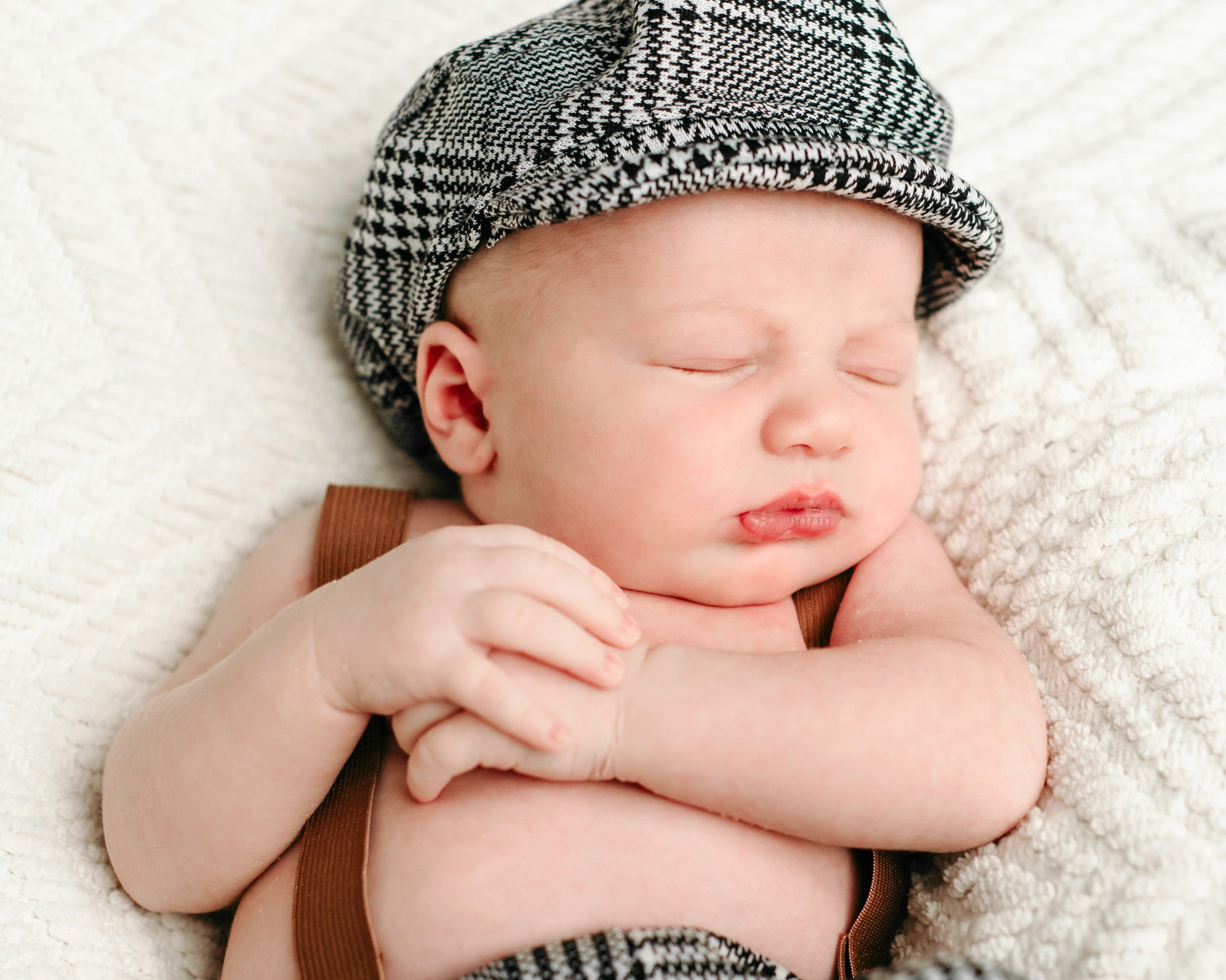 a baby boy wearing a black and white checkered hat and pants with brown suspenders sleeping on a textured white backdrop with his hands tucked up against his chest during a newborn photos session