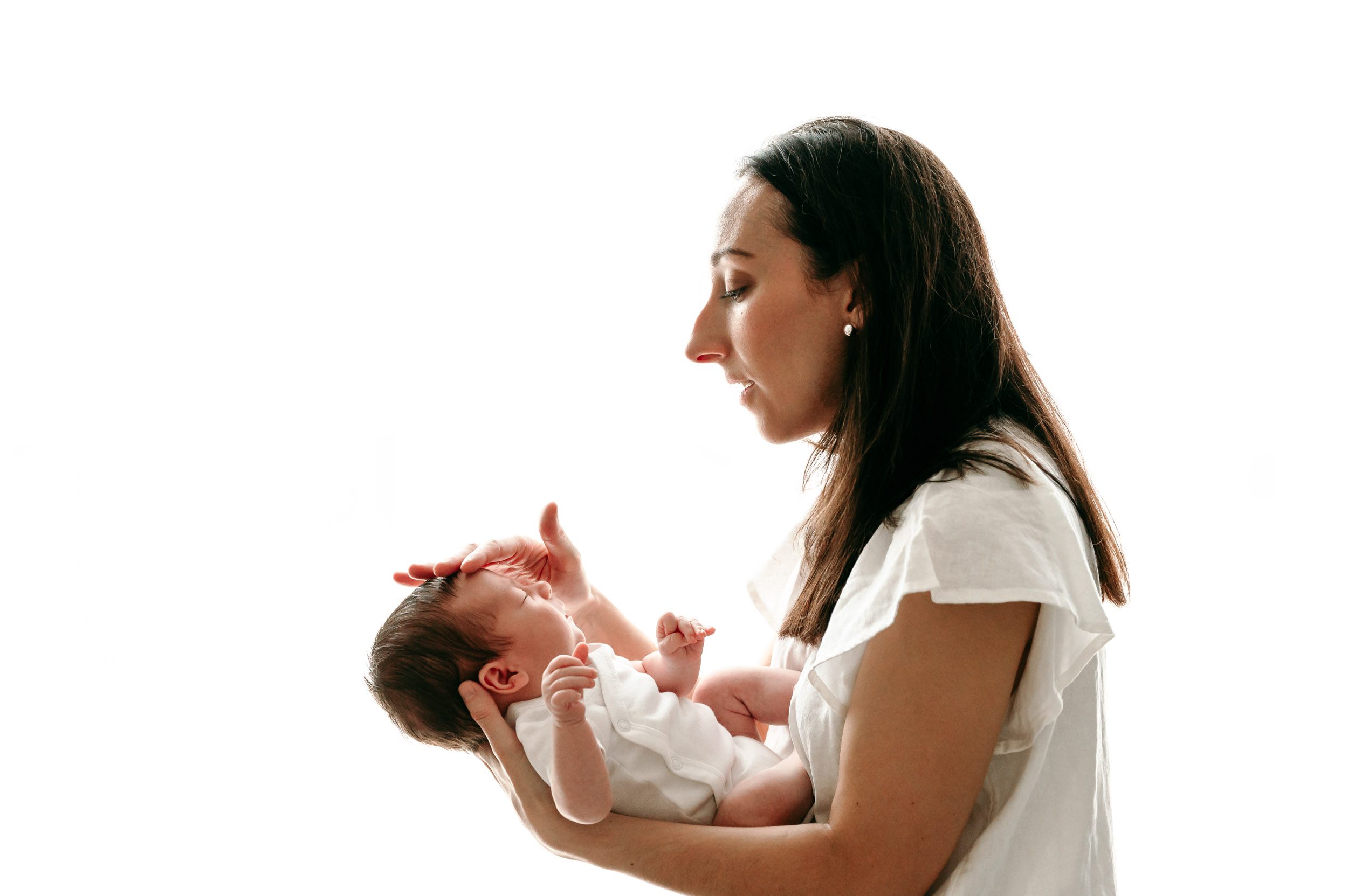 a backlit photo of a new mom wearing a white blouse gently touching her baby boy on the head as she gazes down at him adoringly during a newborn photos session