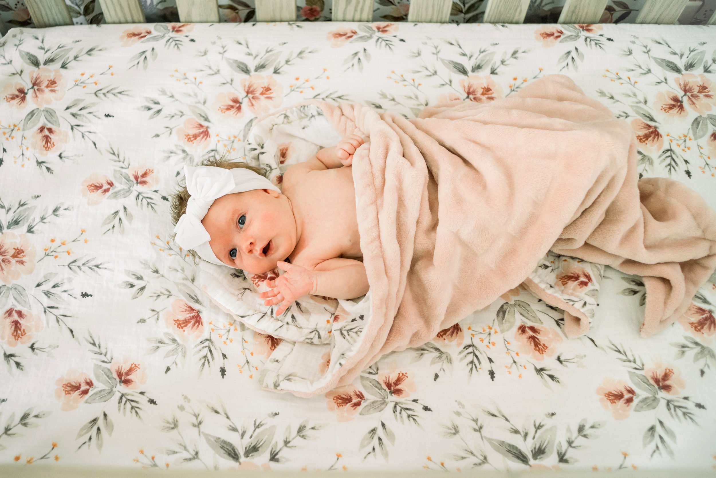 a baby girl wearing a white headband laying in her crib on a floral crib sheet with a fuzzy pink blanket over her gazing up and holding her hand out toward the camera during a newborn photo session