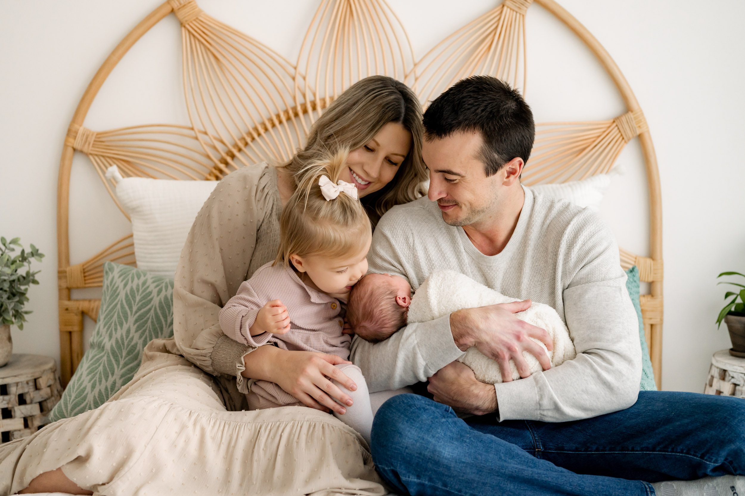 parents sitting on a bed with their baby girl in dad's arms and their older daughter sitting on mom's lapping and kissing her baby sister on the head during a newborn photography session