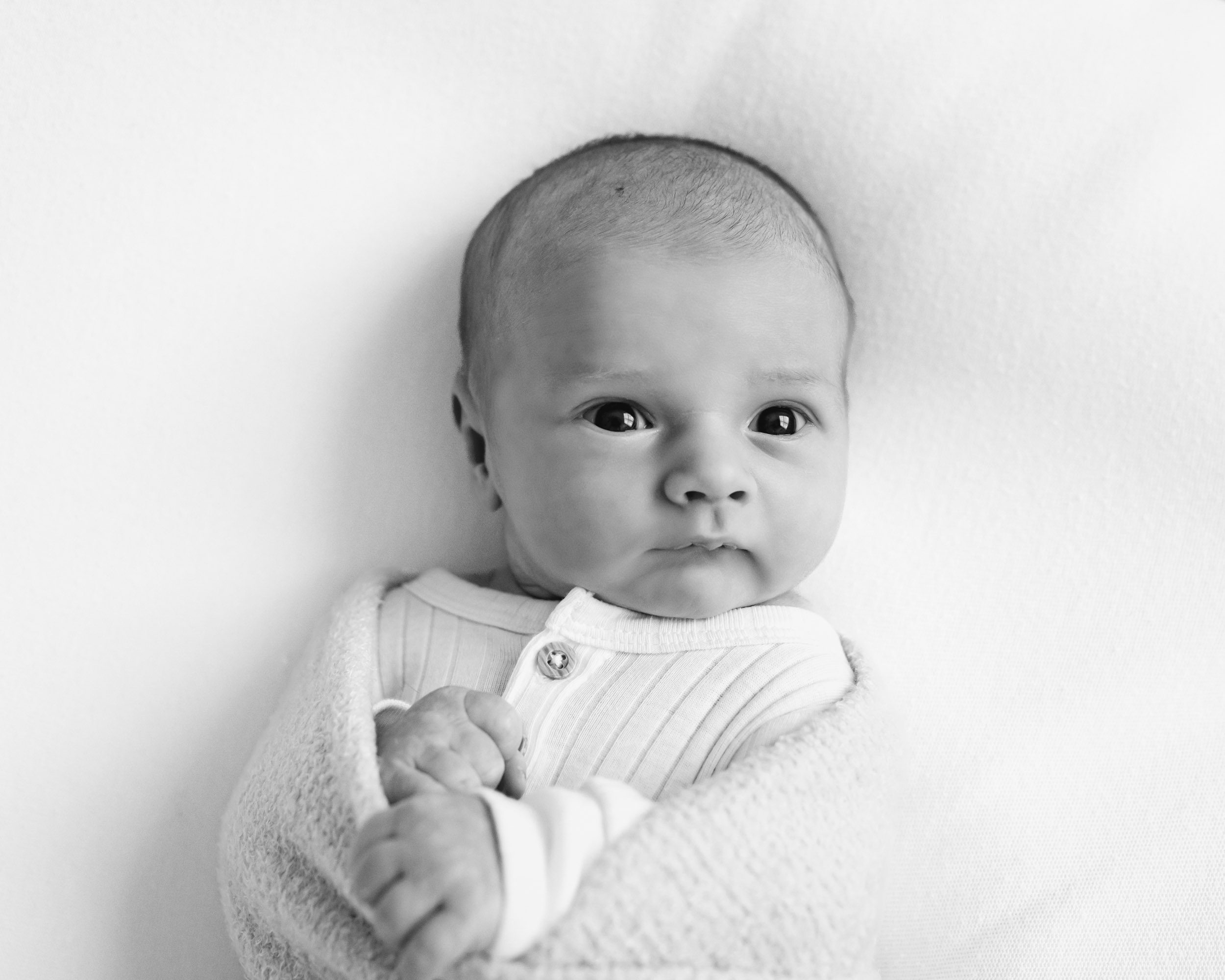 a black and white picture of a newborn baby boy in a white romper and swaddle looking toward the camera with a pensive expression during a newborn baby photography session