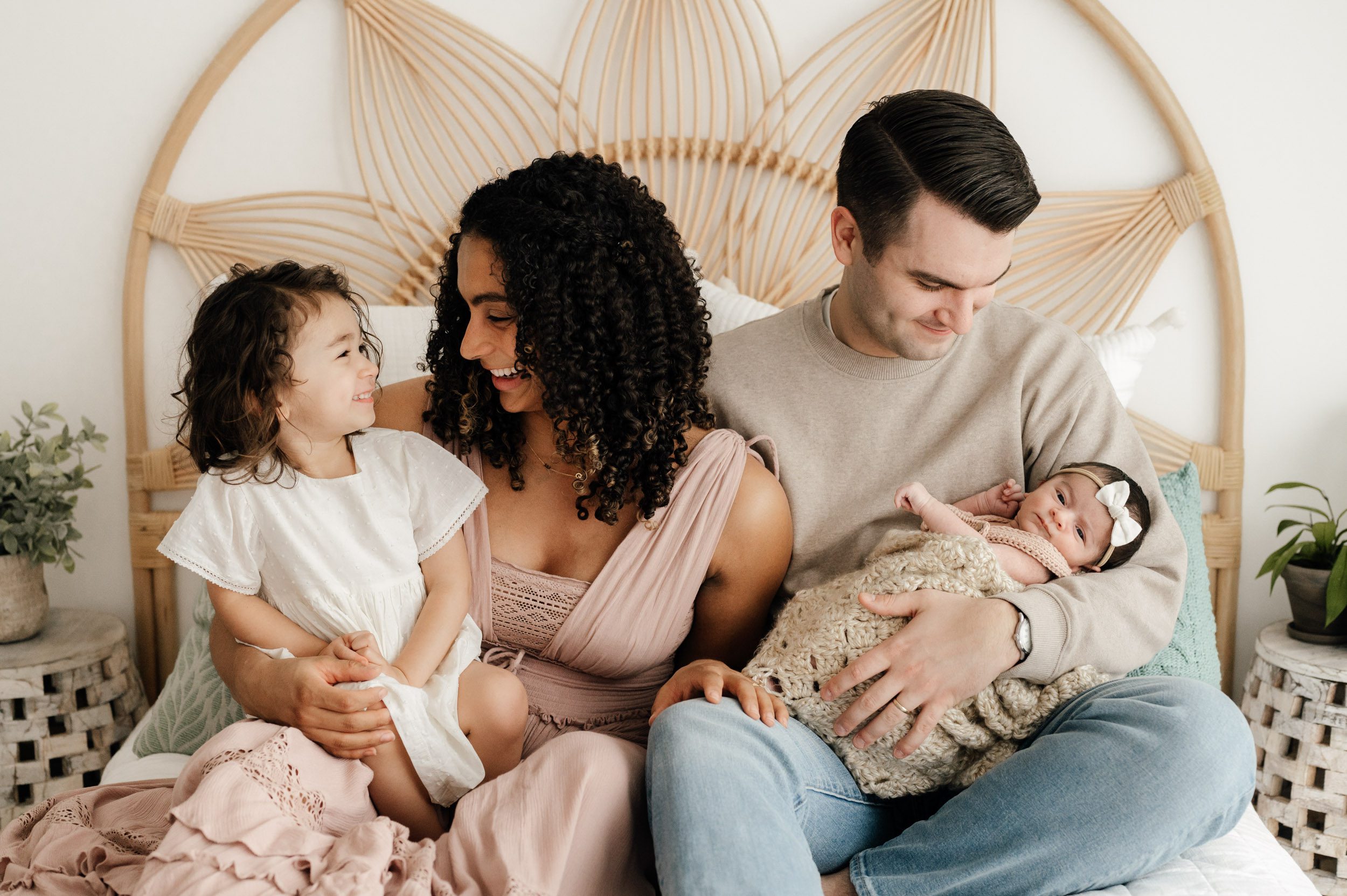 a family of four sitting on a bed with mom smiling at her older daughter and dad smiling down at his baby girl in his arms during a newborn photos session
