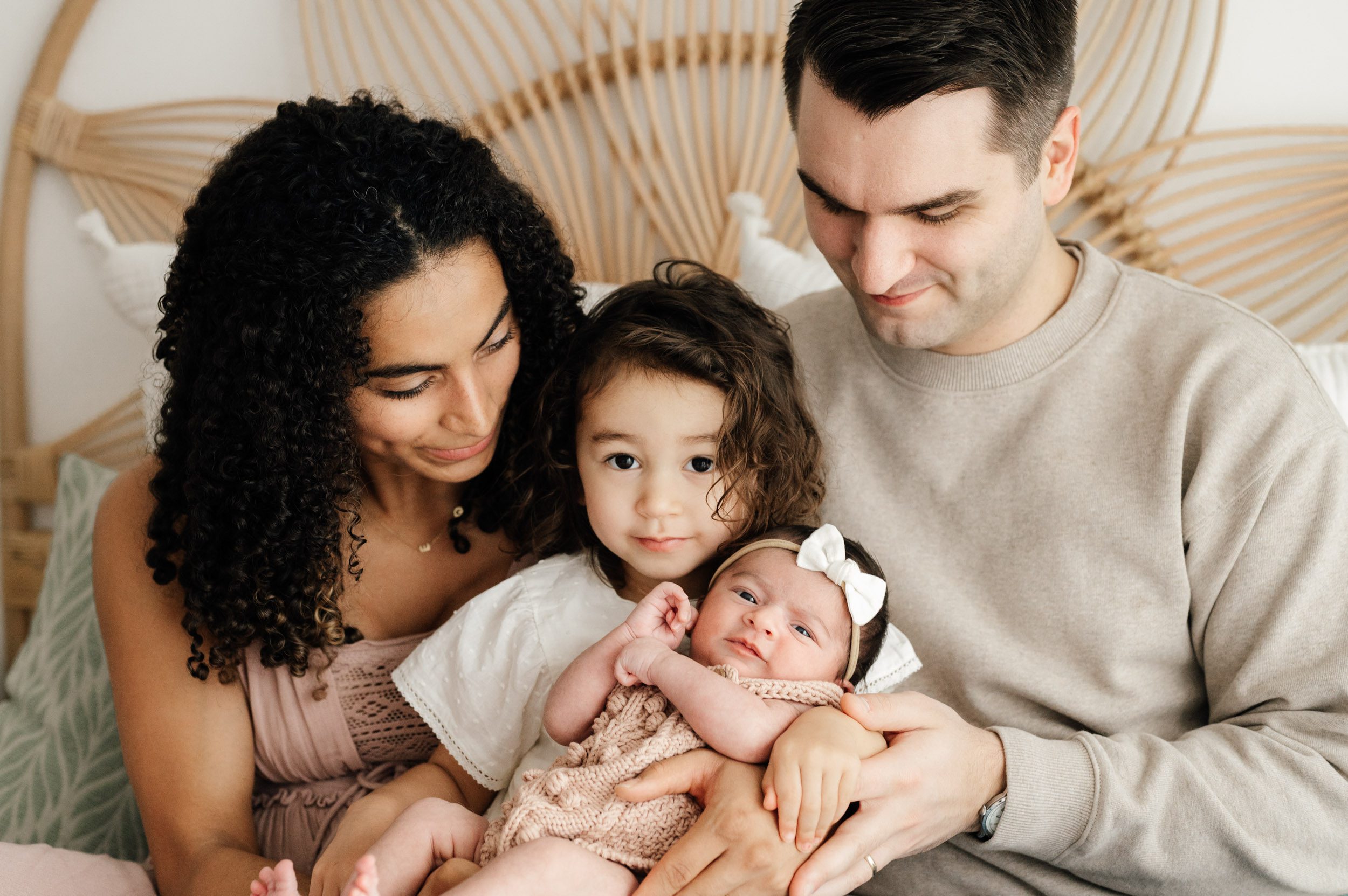 parents sitting on a bed and smiling at their newborn baby girl and older daughter in their lap as big sister and baby girl look directly at the camera during a natural light newborn photo session