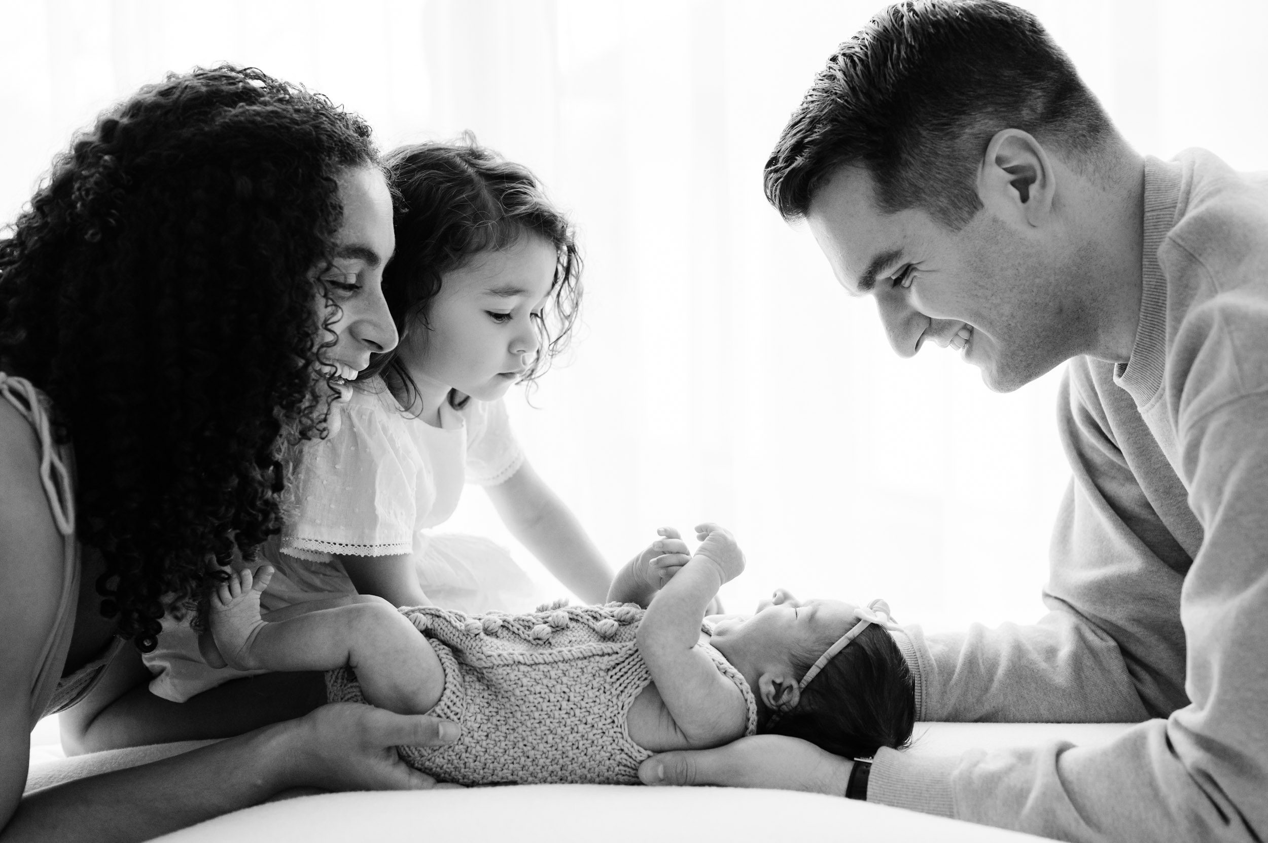 a black and white backlit photo of a newborn baby girl laying on a bean bag surrounded by her parents and older sister during a newborn photoshoot
