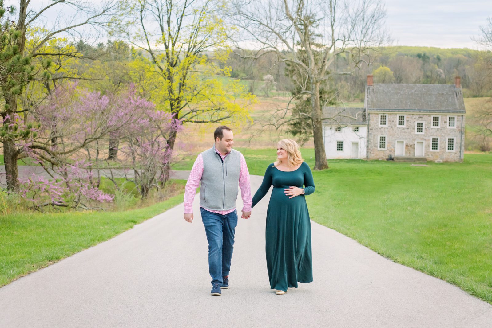 an expecting couple holding hands and smiling at each other as they walk along a pathway with a purple redbud tree blooming in the background during a spring photos session