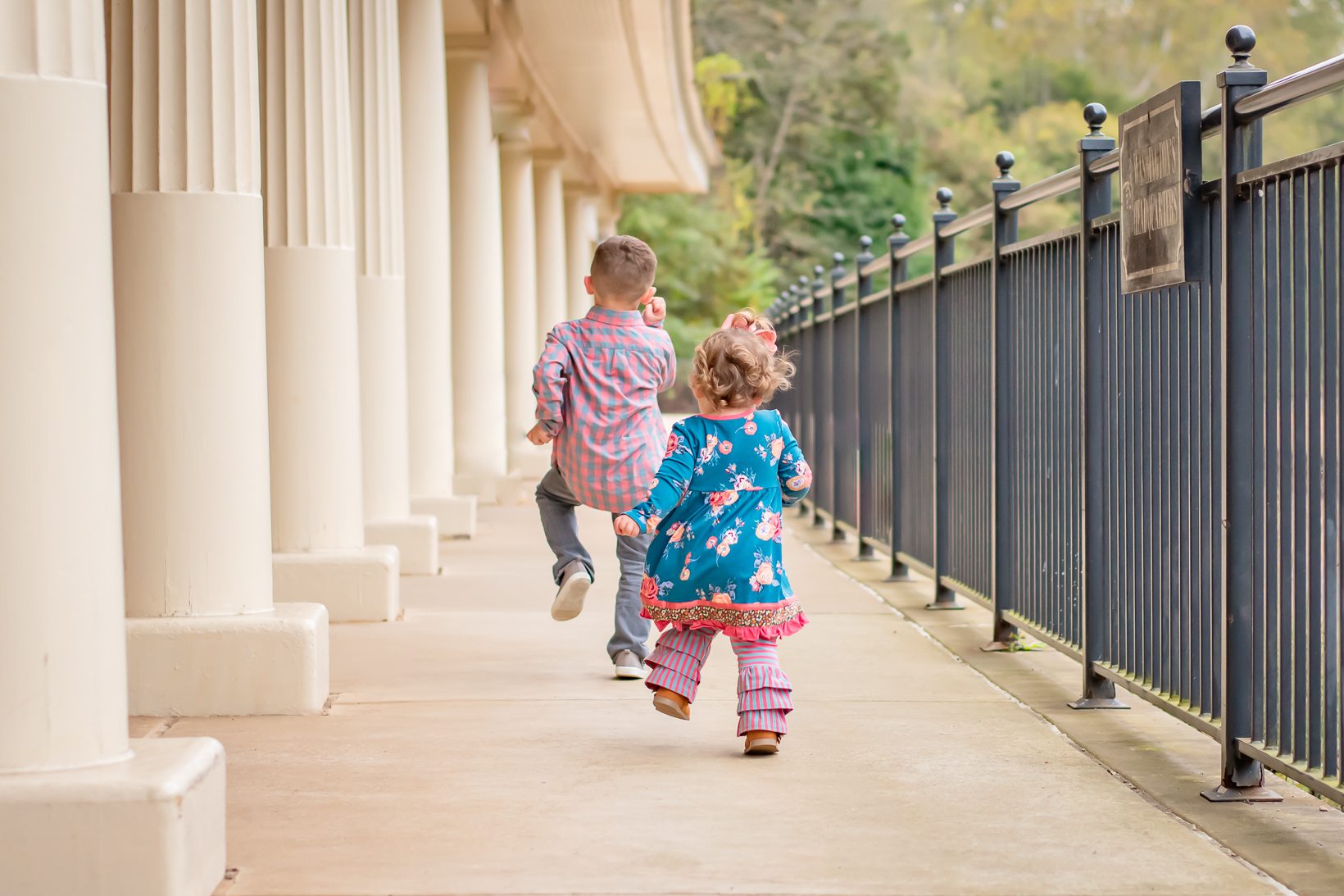 two young kids marching down a walkway with a line of white columns on one side and a black slatted fence on the other side during a spring photo session