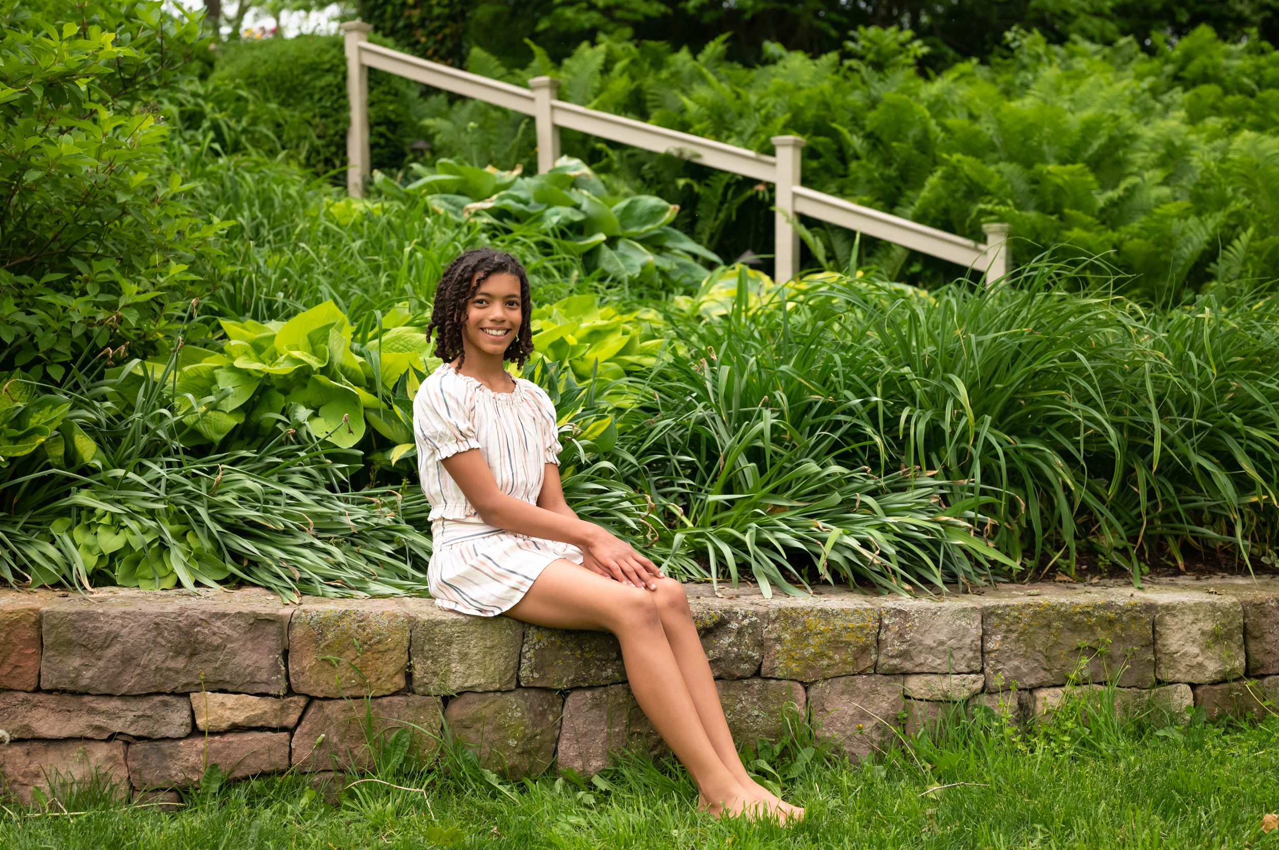 a girl sitting on a stone wall with a garden in the background and smiling at the camera during a family photoshoot