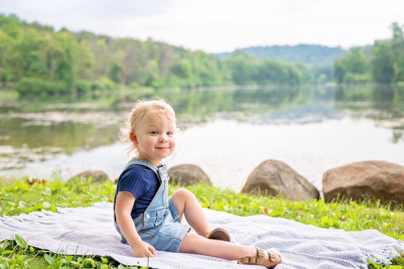 a little boy sitting on a blanket next to a lake and smiling at the camera during a spring photos session