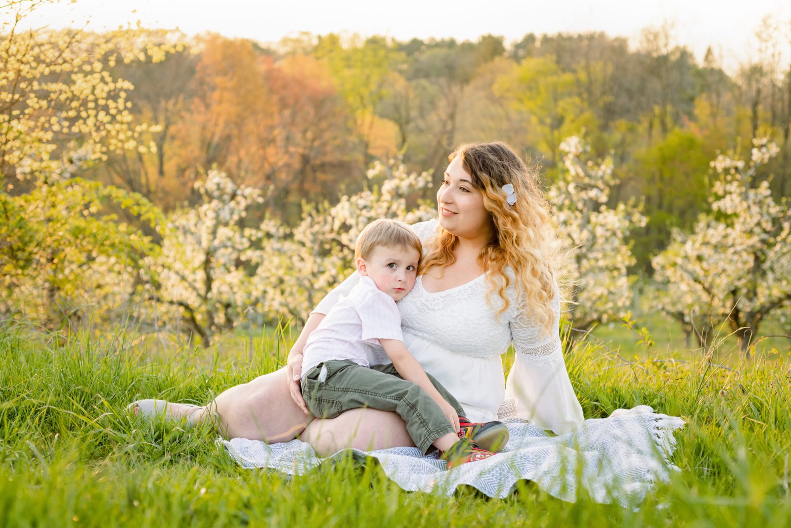 a mom sitting on a blanket in an orchard full of apple trees covered in white blossoms and snuggling with her son on her lap as she smiles off into the distance during a spring photo session