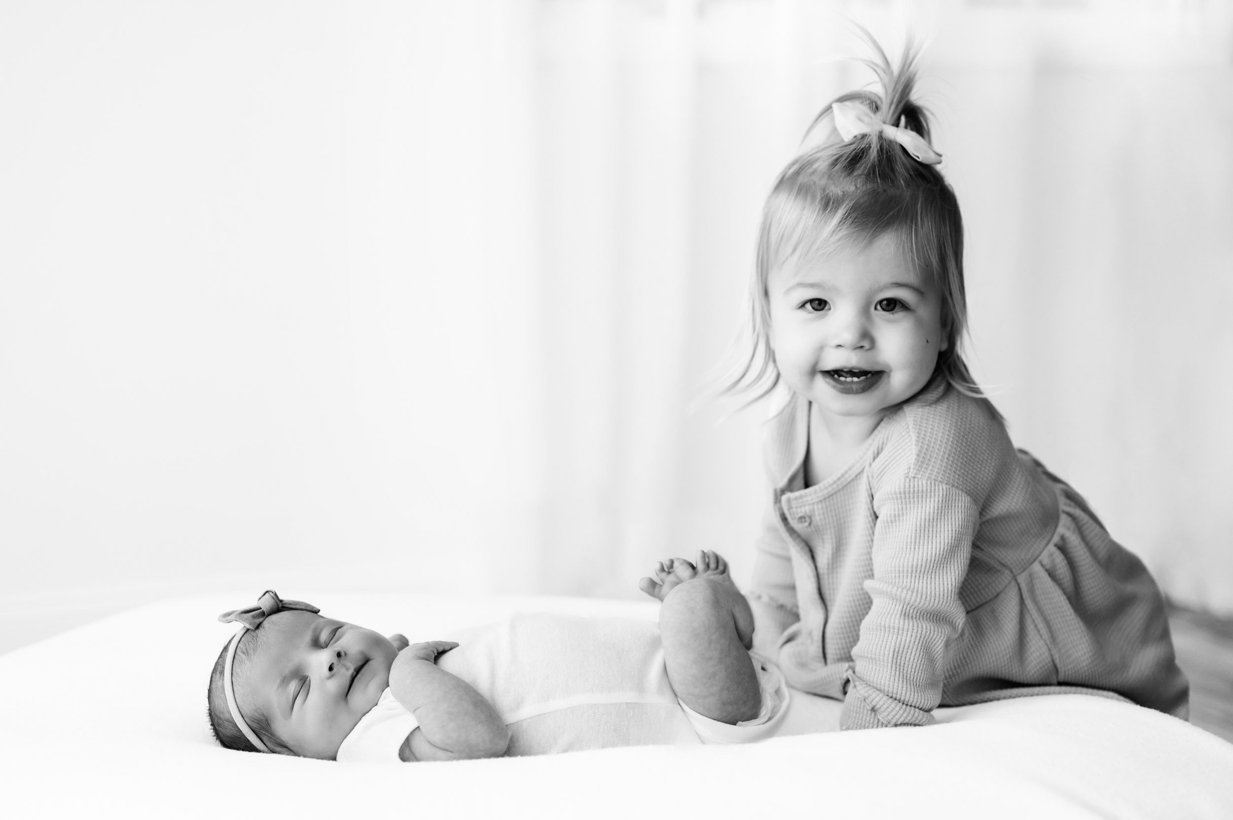 a black and white picture of a baby girl laying on a white bean bag while her older sister stands next to her and smiles at the camera during a Chester County newborn photo session