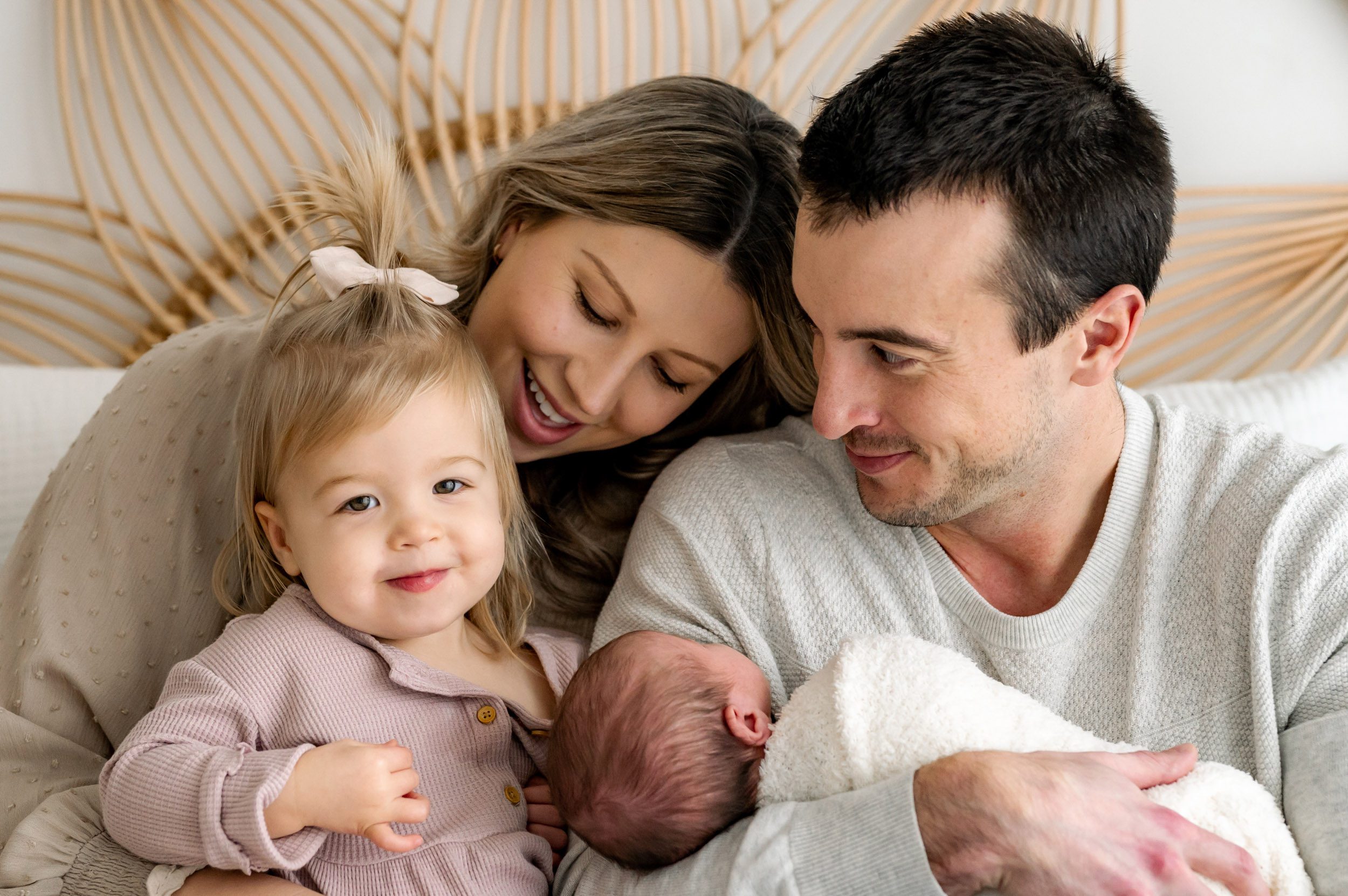 a mom and dad sitting on a bed and snuggling with their baby girl and older daughter on their lap as the older daughter looks at the camera and smiles during a Chester County newborn photography session