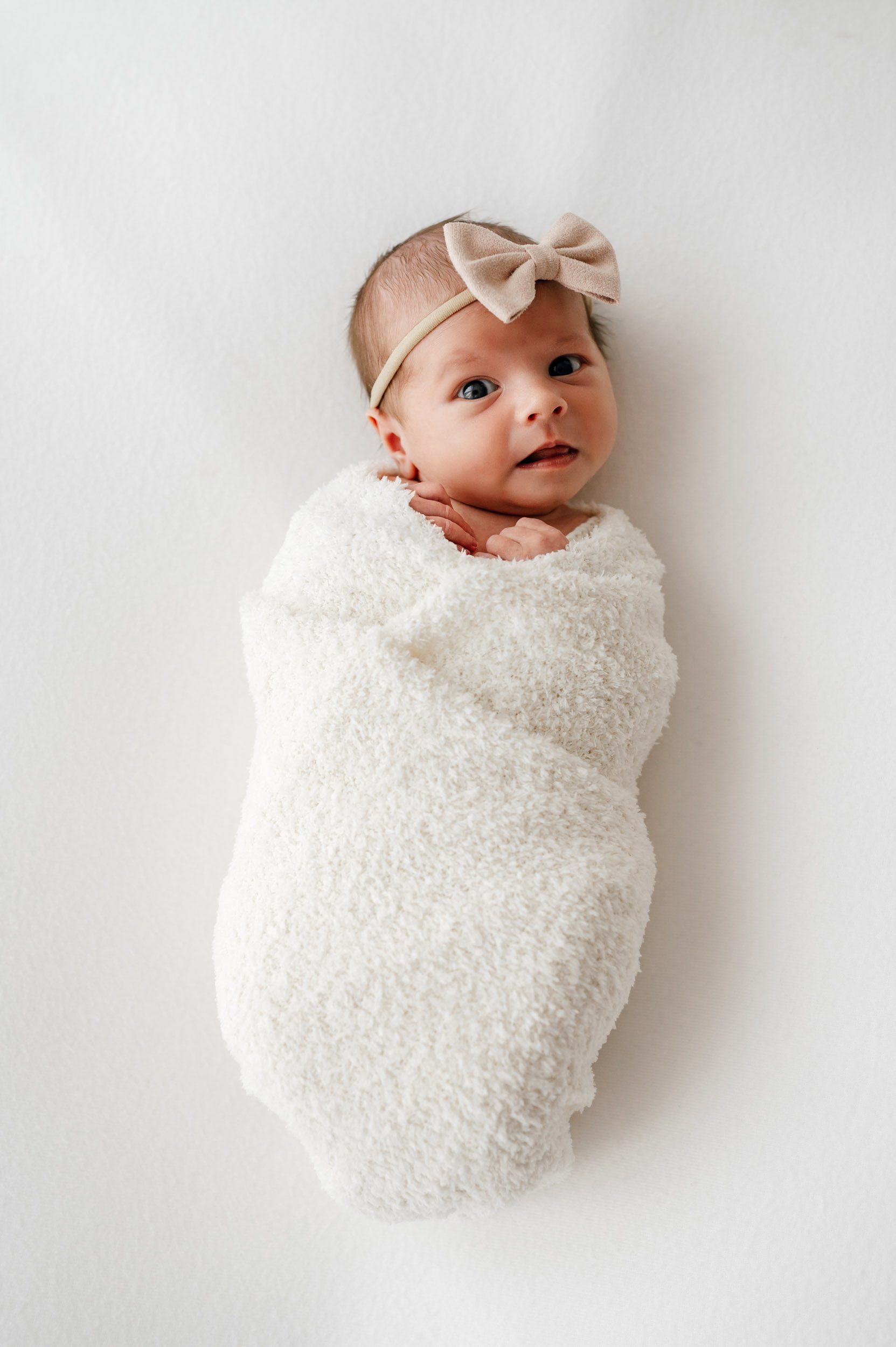 a baby girl wrapped in a fuzzy white swaddle wearing a headband with a light pink bow gazing up at the camera during a newborn photography session