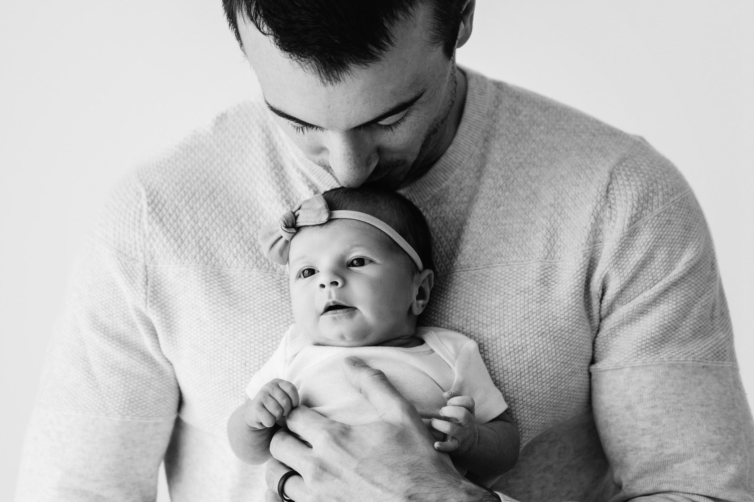a black and white picture of a new dad holding his baby girl against his chest and kissing her on top of her head as she looks toward the window light during a Chester County newborn photography session