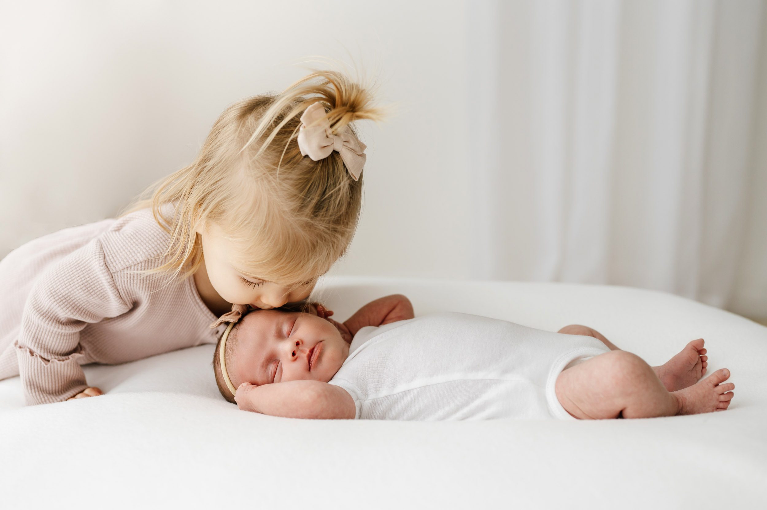 a baby girl laying on a white bean bag while her older sister gently kisses her on the head during a newborn photoshoot