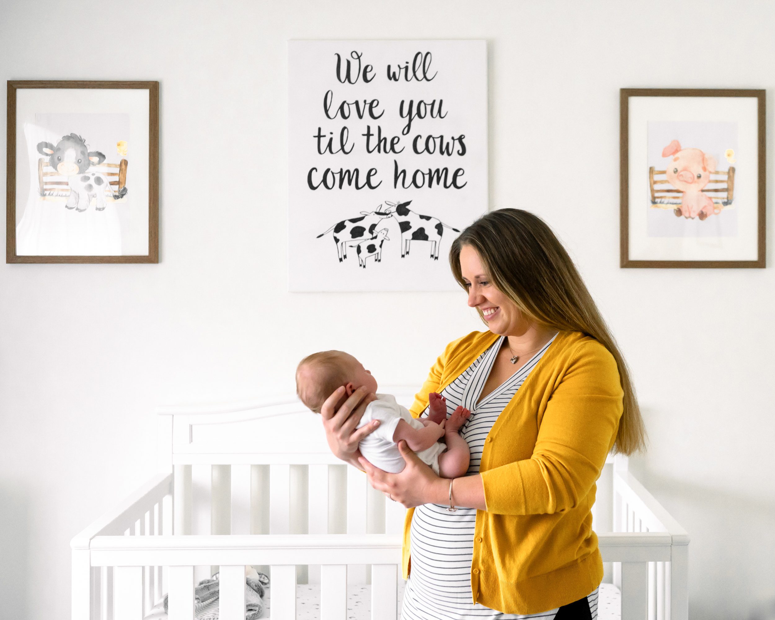 a new mom wearing a striped shirt and yellow cardigan standing in front of the crib in her son's nursery and holding him in her arms as she smiles down at him during a newborn photos session