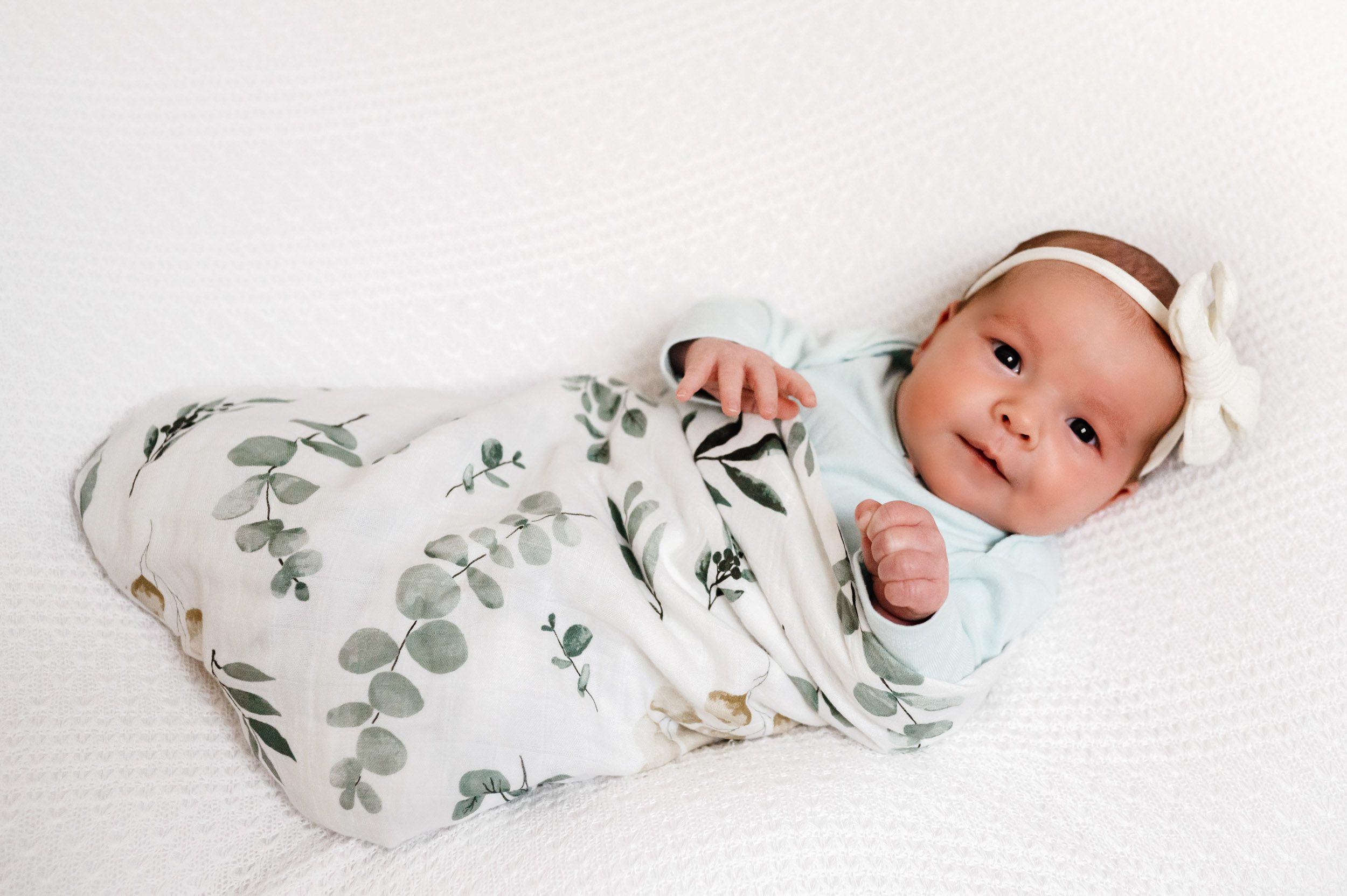 a baby girl in floral swaddle blanket laying on a white textured backdrop looking directly at the camera with a hint of a smile on her face during a newborn photoshoot