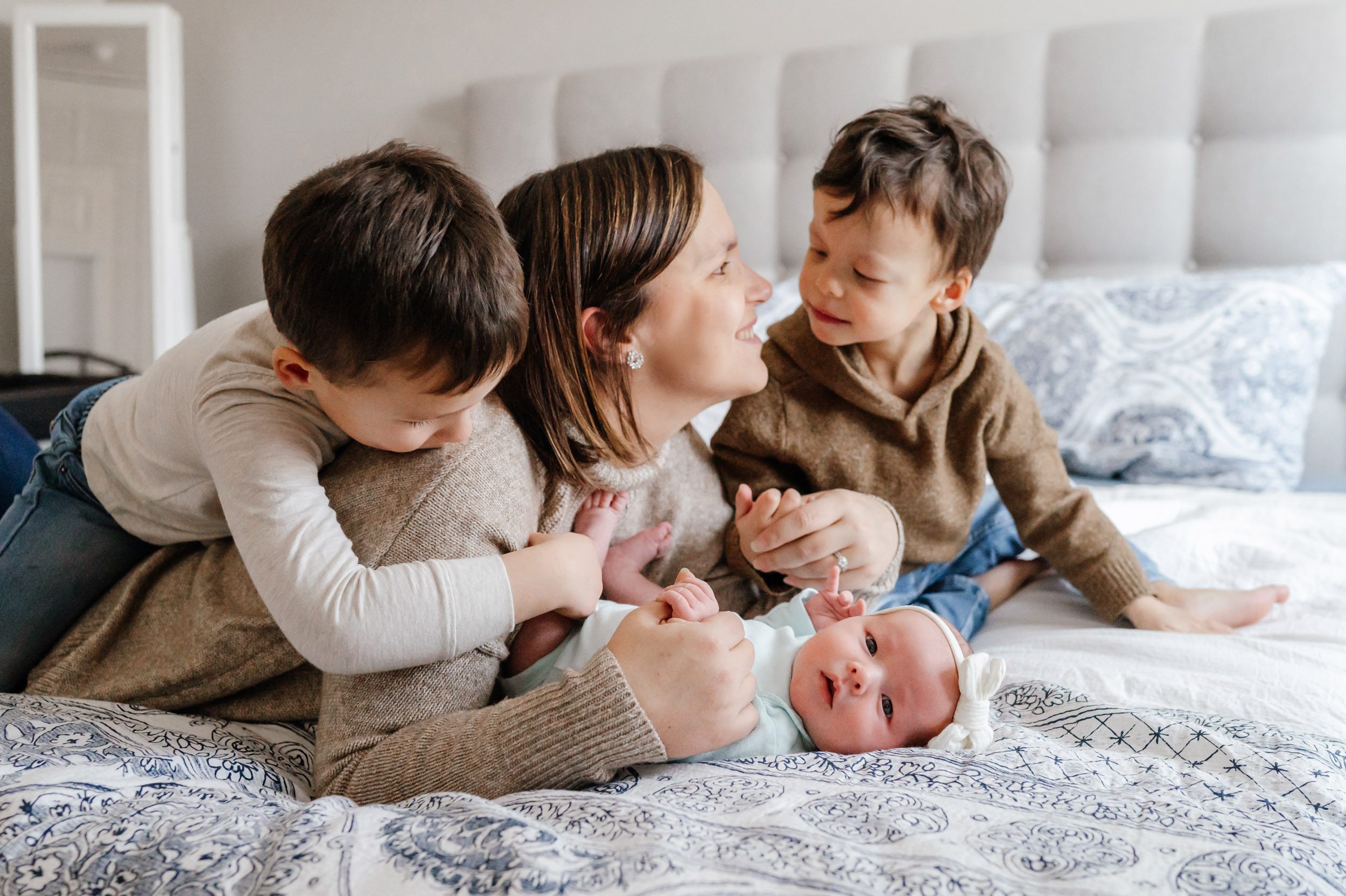 a mom laying on a bed with her oldest son laying on her back and hugging her, her younger son looking at her and smiling, and her baby girl laying in front of her and looking at the camera during an Exton newborn photo session