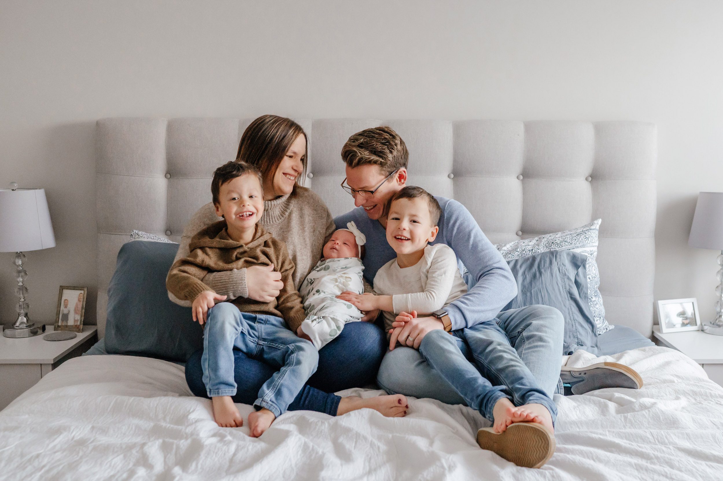 a mom and dad sitting on their bed with their baby girl and two older sons snuggled in their laps as everyone laughs during an Exton in home newborn session