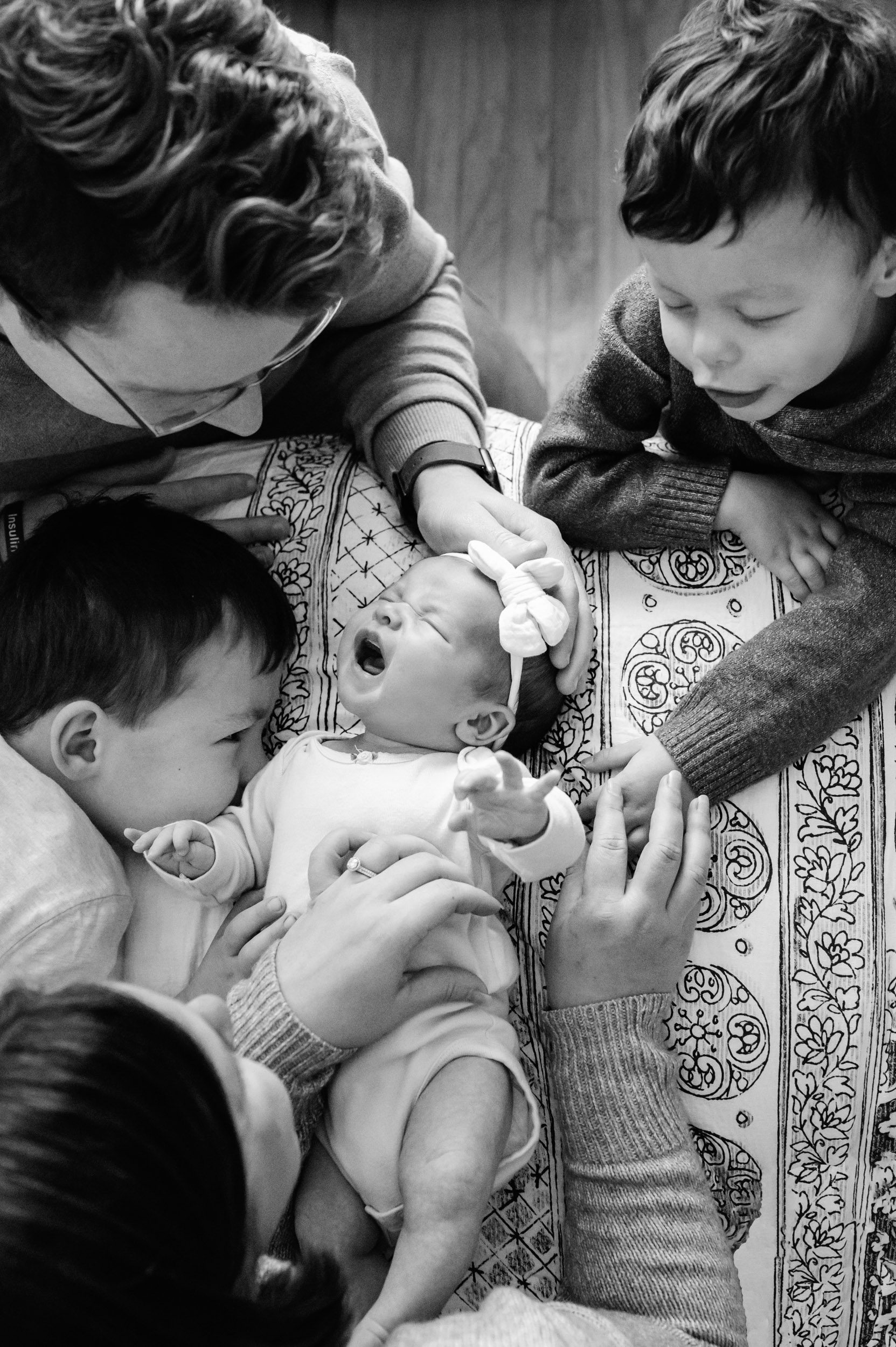 a black and white picture taken from above of a baby girl laying on a bed and crying while her parents and two older brothers surround her and comfort her by touching her with their hands during an Exton newborn photography session