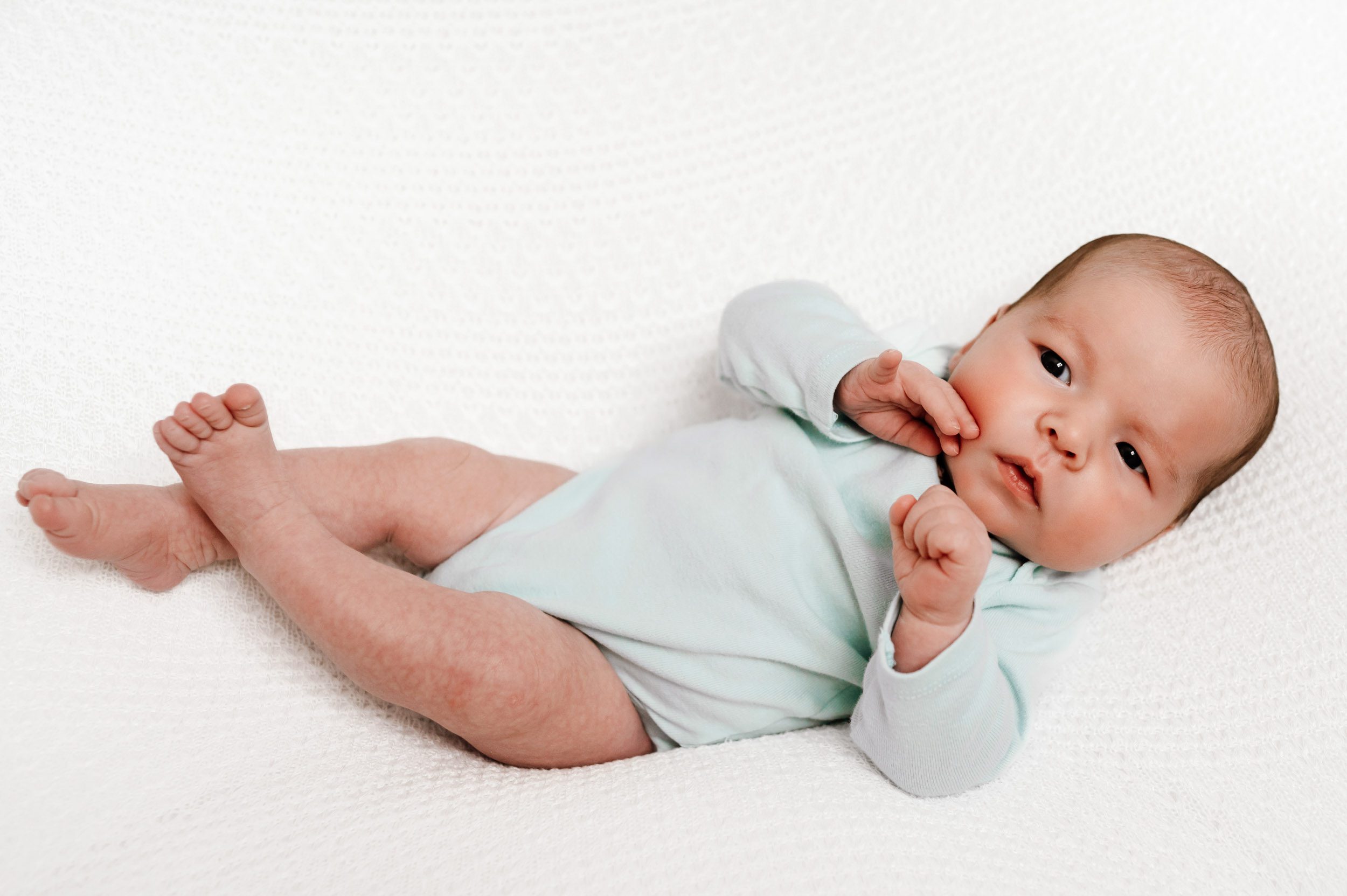 a baby girl laying on a white textured backdrop and touching her cheek during a newborn photoshoot