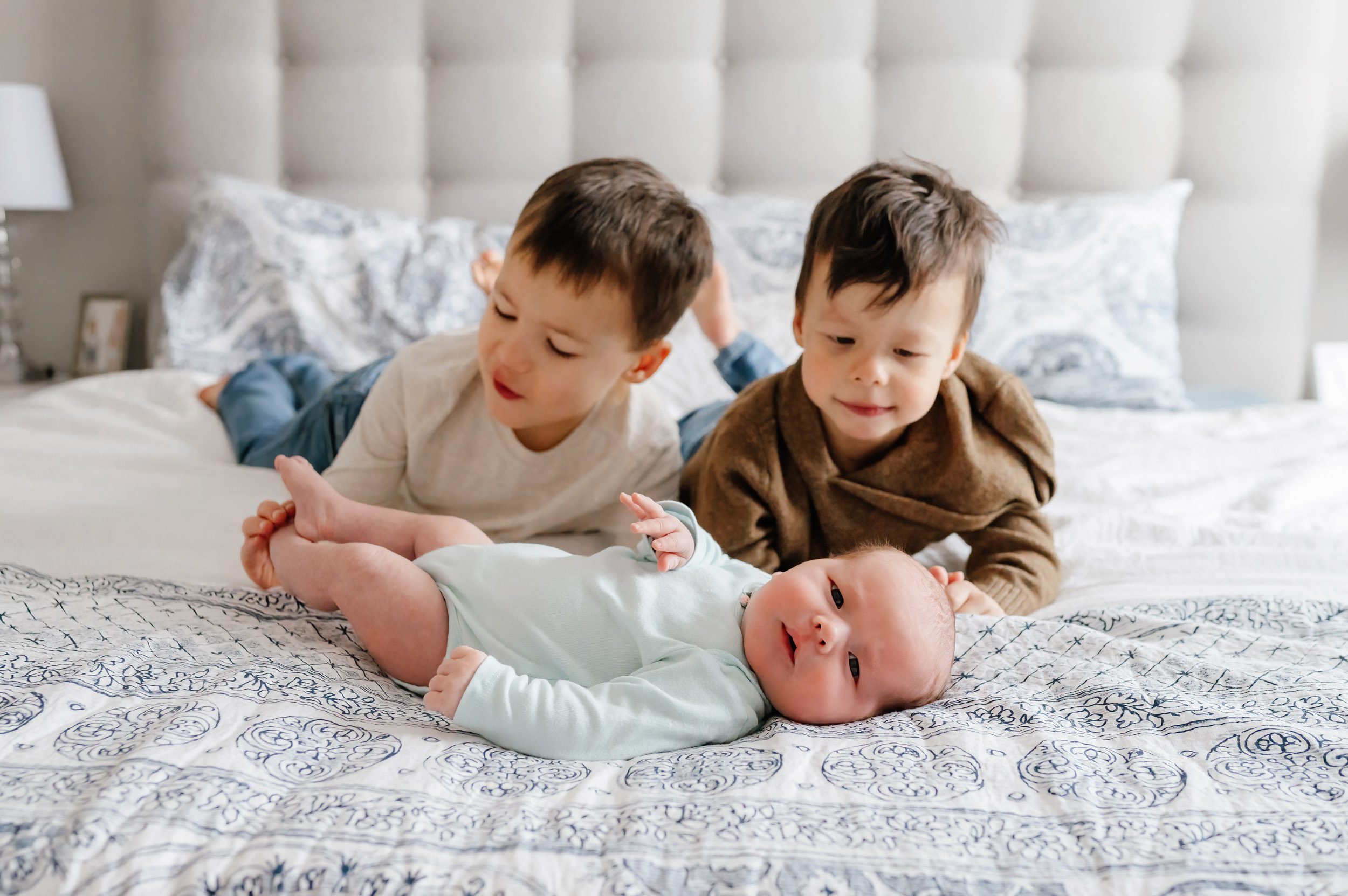 a baby girl laying on a bed and looking at the camera while her two older brothers lay behind her and touch her toes and smile at her during an Exton newborn photography session