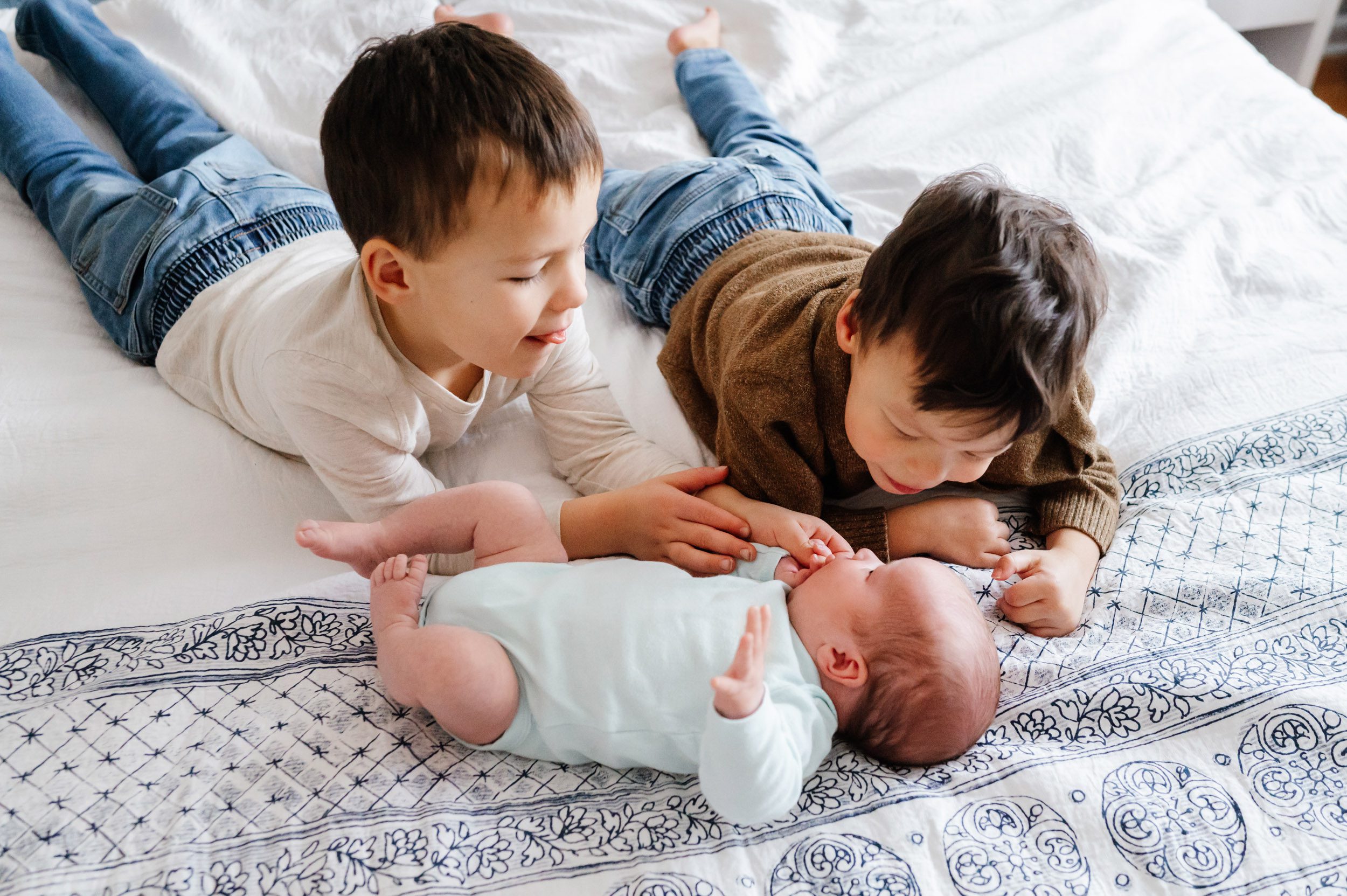 a baby girl laying on a bed while her two older brothers lay next to her and touch her hands as they smile down at her during an Exton newborn photography session