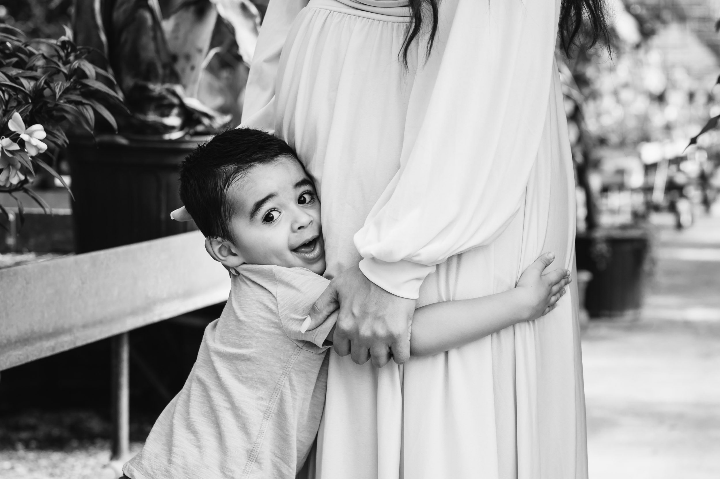 a black and white picture of a young boy hugging his expecting mom's belly and looking toward the camera with a huge grin on his face during a Collegeville maternity photography session