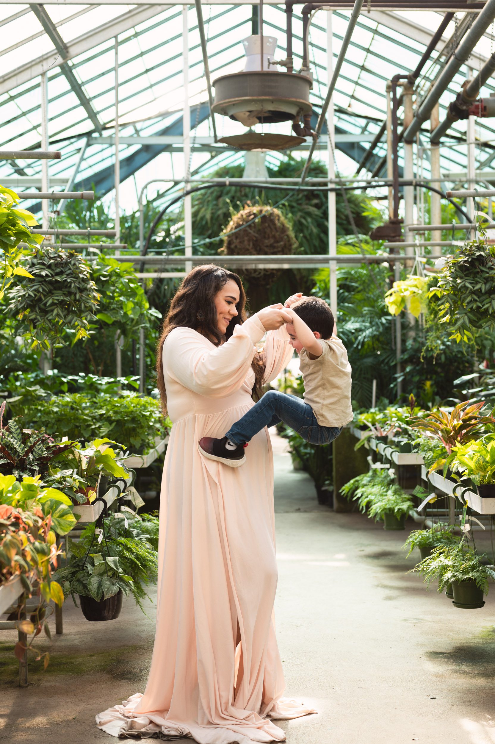 an expecting mom standing in a greenhouse and lifting her young son in the air with her arms during a maternity photoshoot