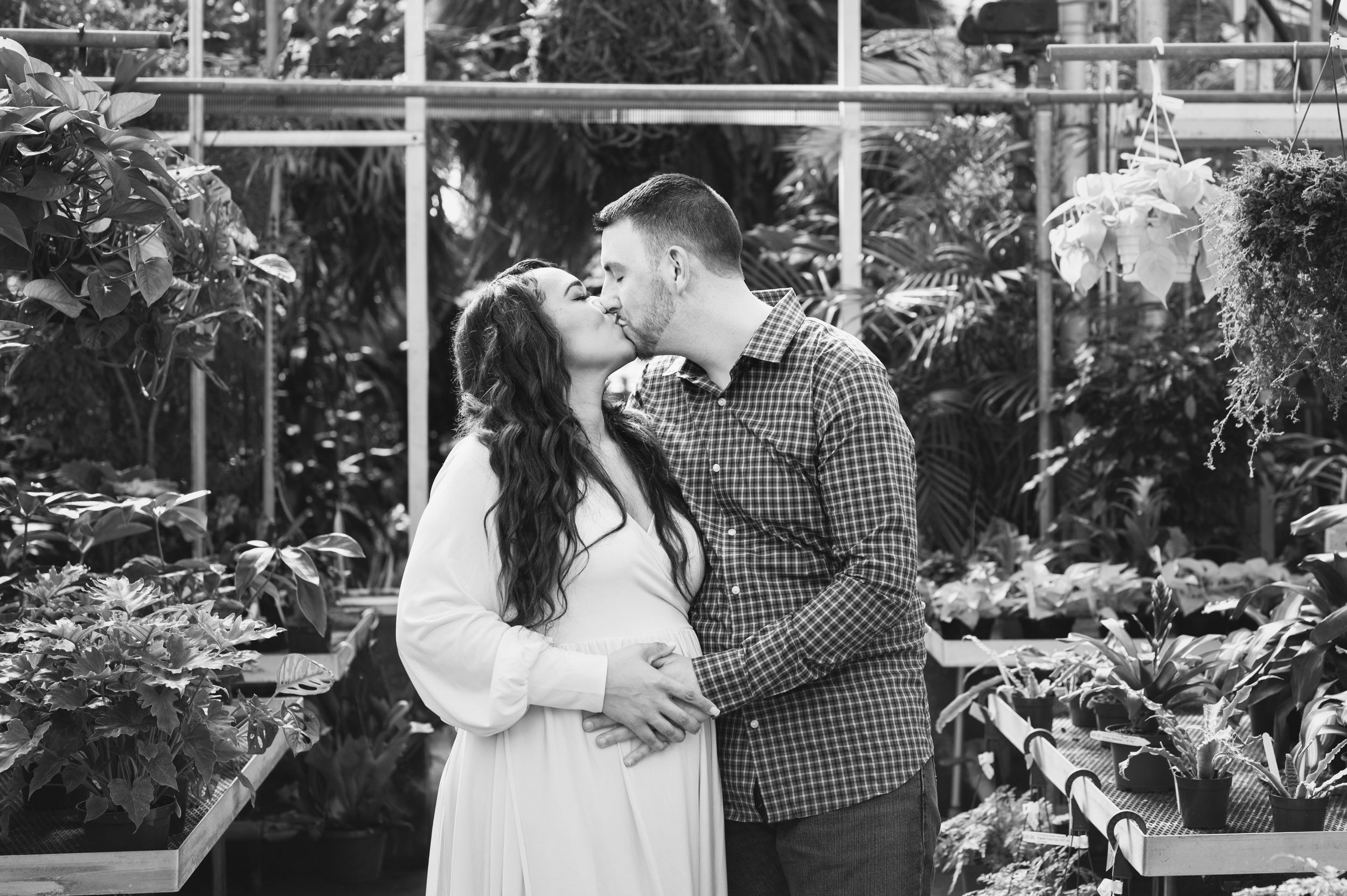 a black and white picture of expecting parents standing in a greenhouse and both touching mom's belly and kissing each other during a maternity photoshoot