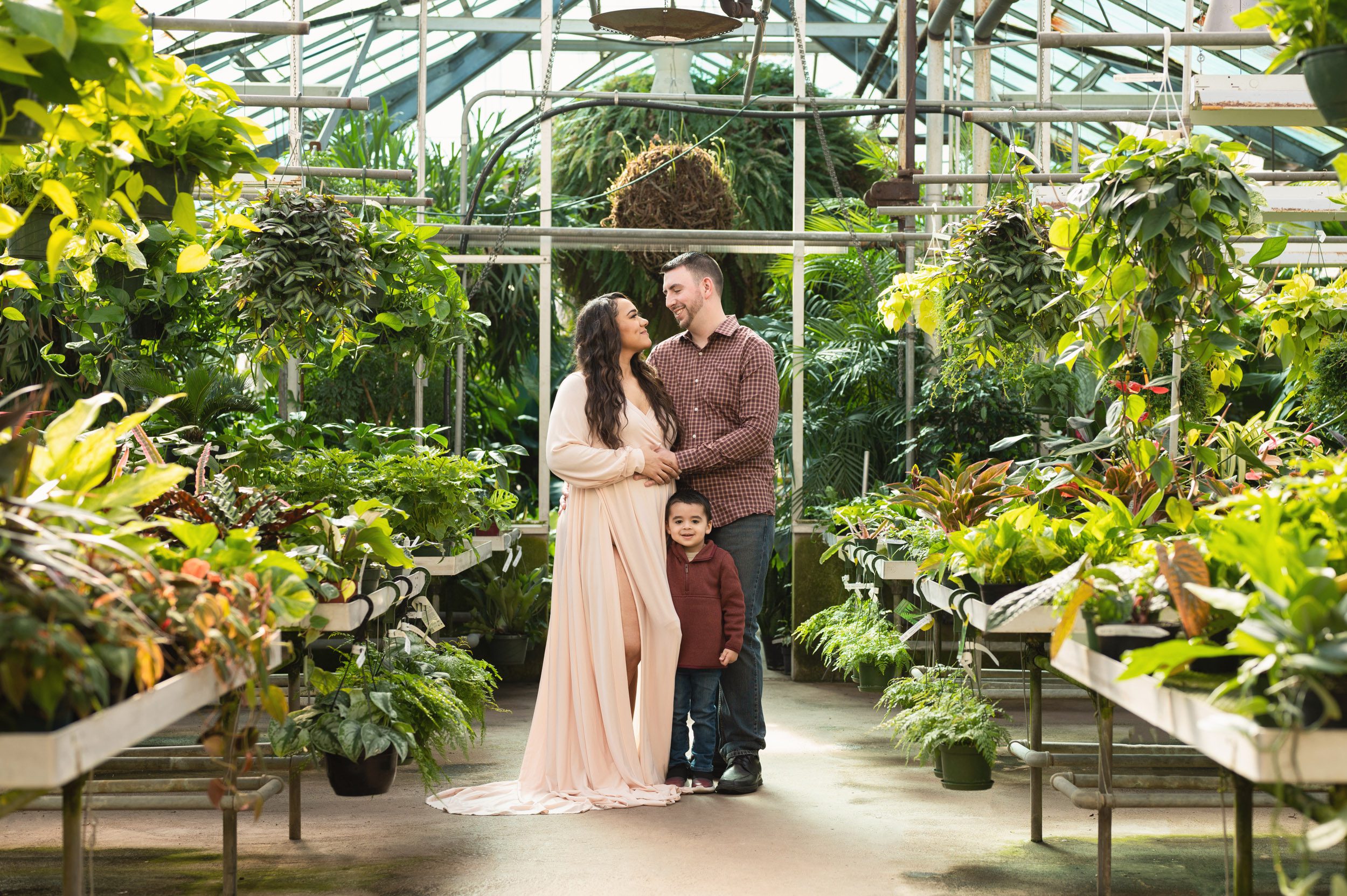 expecting parents standing in a greenhouse and smiling at each other with their young son hugging mom's leg and smiling at the camera during a Collegeville greenhouse maternity photography session