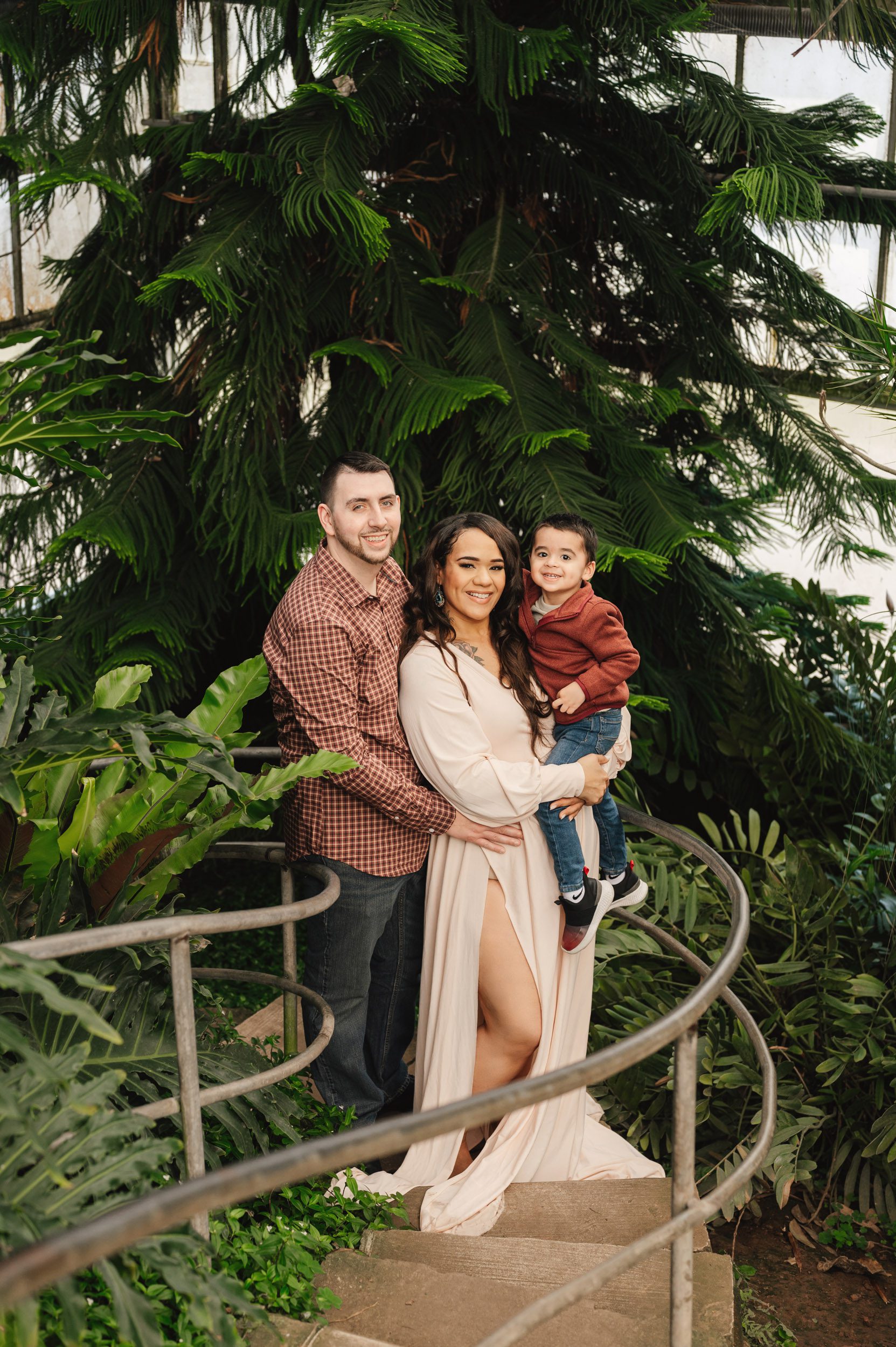 a family of three standing on curved staircase in a greenhouse smiling at the camera during a maternity photography session