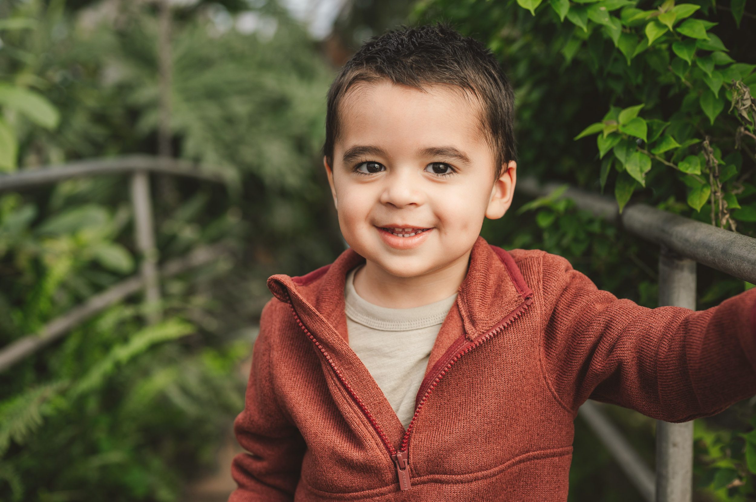 a young boy standing in a greenhouse holding onto a metal railing and smiling directly at the camera during a family maternity photoshoot