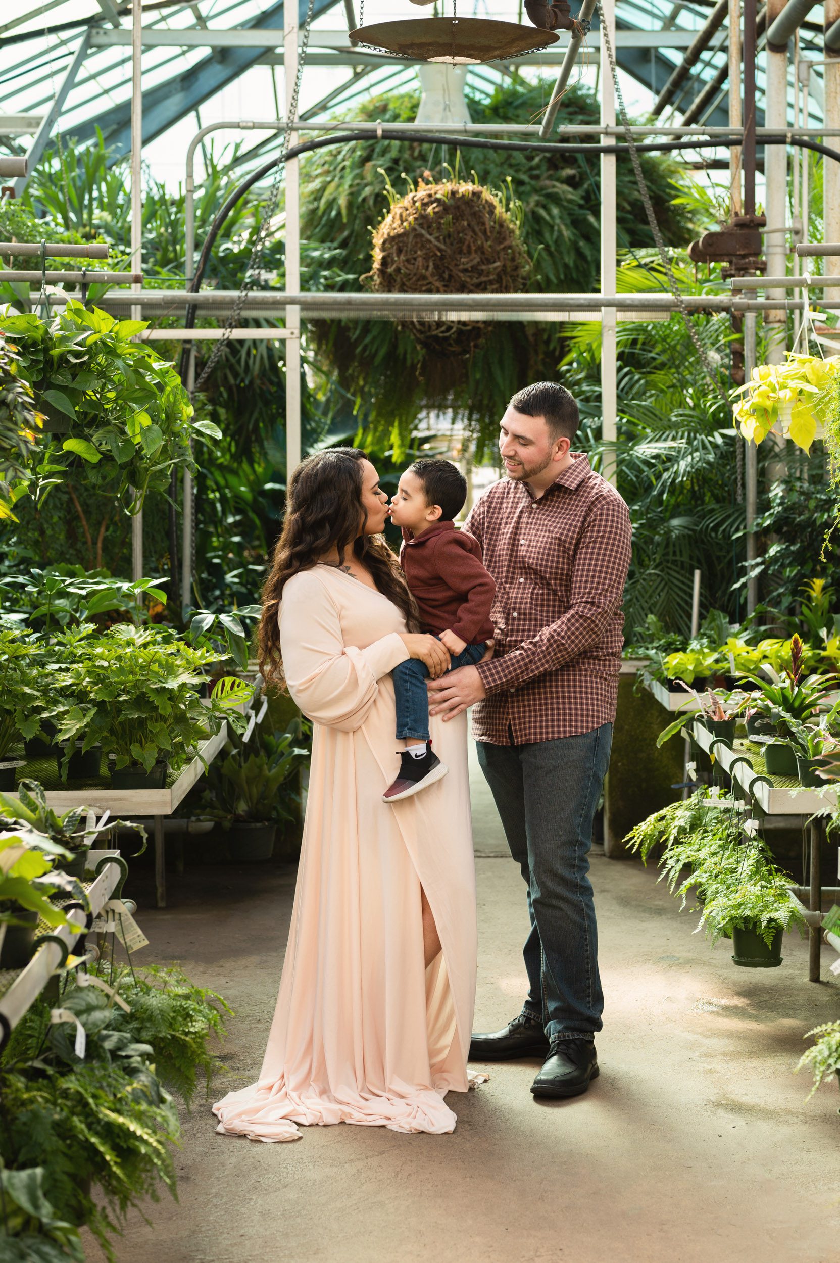 an expecting mom holding her young son on her belly as he gives her a kiss on the lips and dad hugs them and smiles at their son during a greenhouse maternity photoshoot