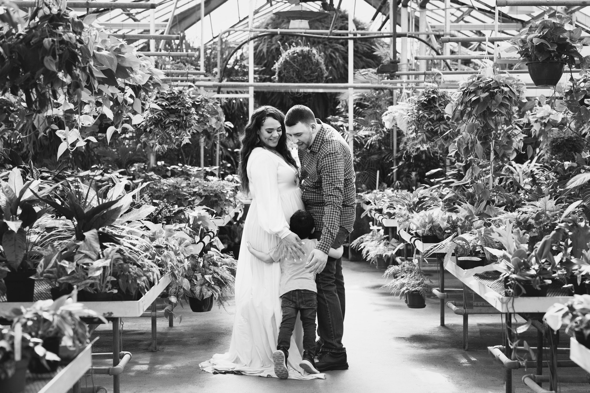 a black and white picture of an expecting mom and dad standing in a greenhouse as their young son runs up and gives them a hug during a Collegeville greenhouse maternity photoshoot