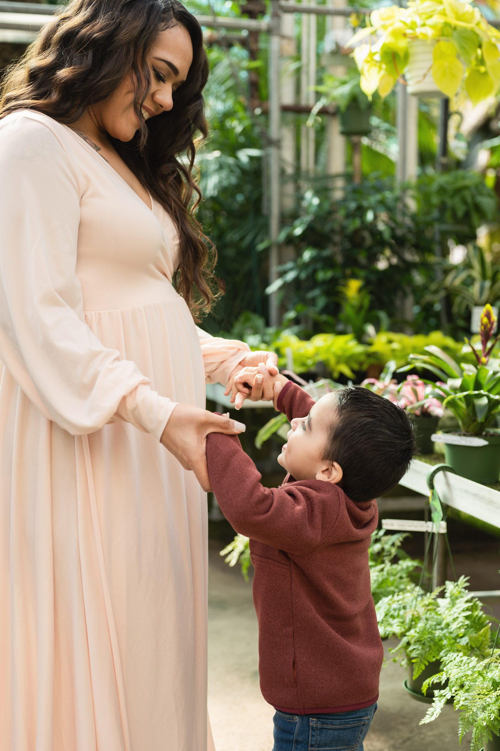 an expecting mom holding her young son's hands and smiling down at him as he looks at her belly during a maternity photoshoot