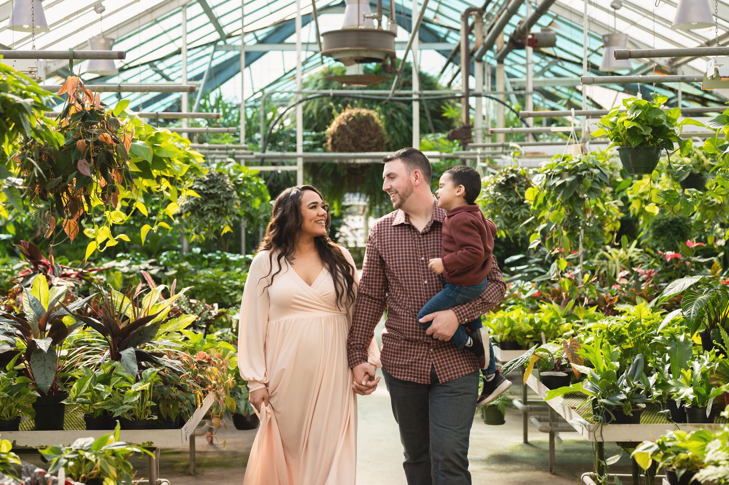 expecting parents holding hands and smiling at each other as they walk through a greenhouse while dad holds their young son on his hip during a Collegeville maternity photography session