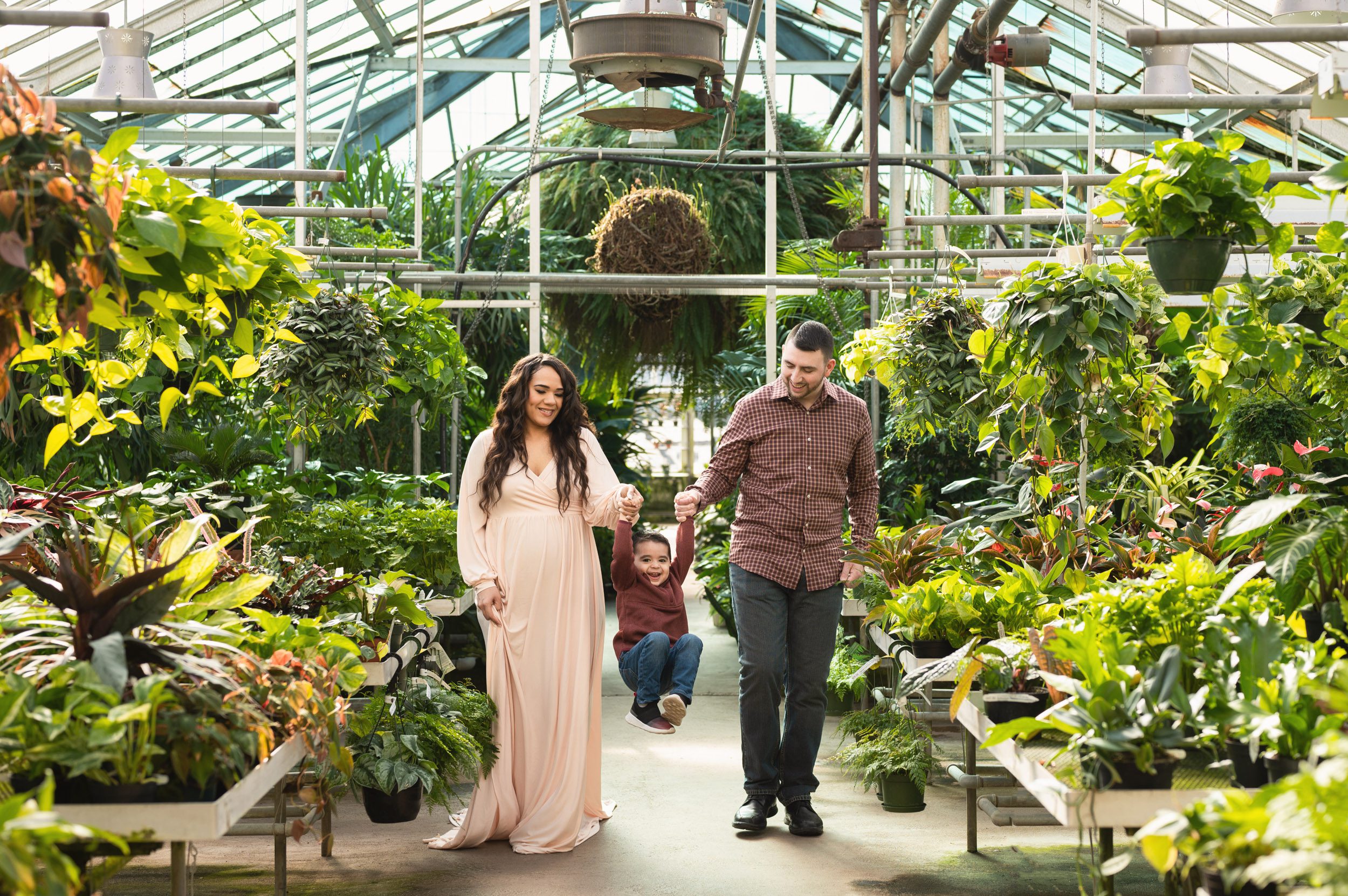 a young boy holding his parents hands and swinging up in the air with a huge smile on his face as they walk through a greenhouse during a maternity photography session