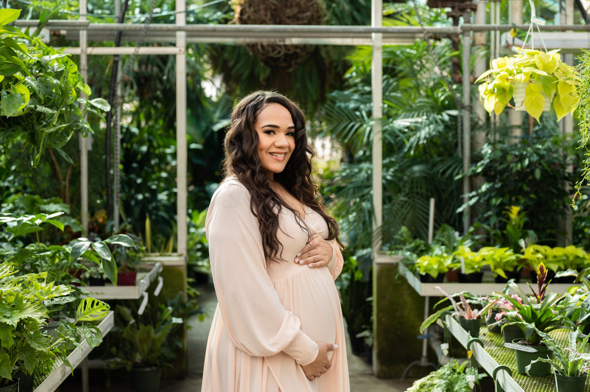 an expecting mom in a pale pink dress standing in a greenhouse and cradling her belly as she smiles at the camera during a Collegeville maternity photography session
