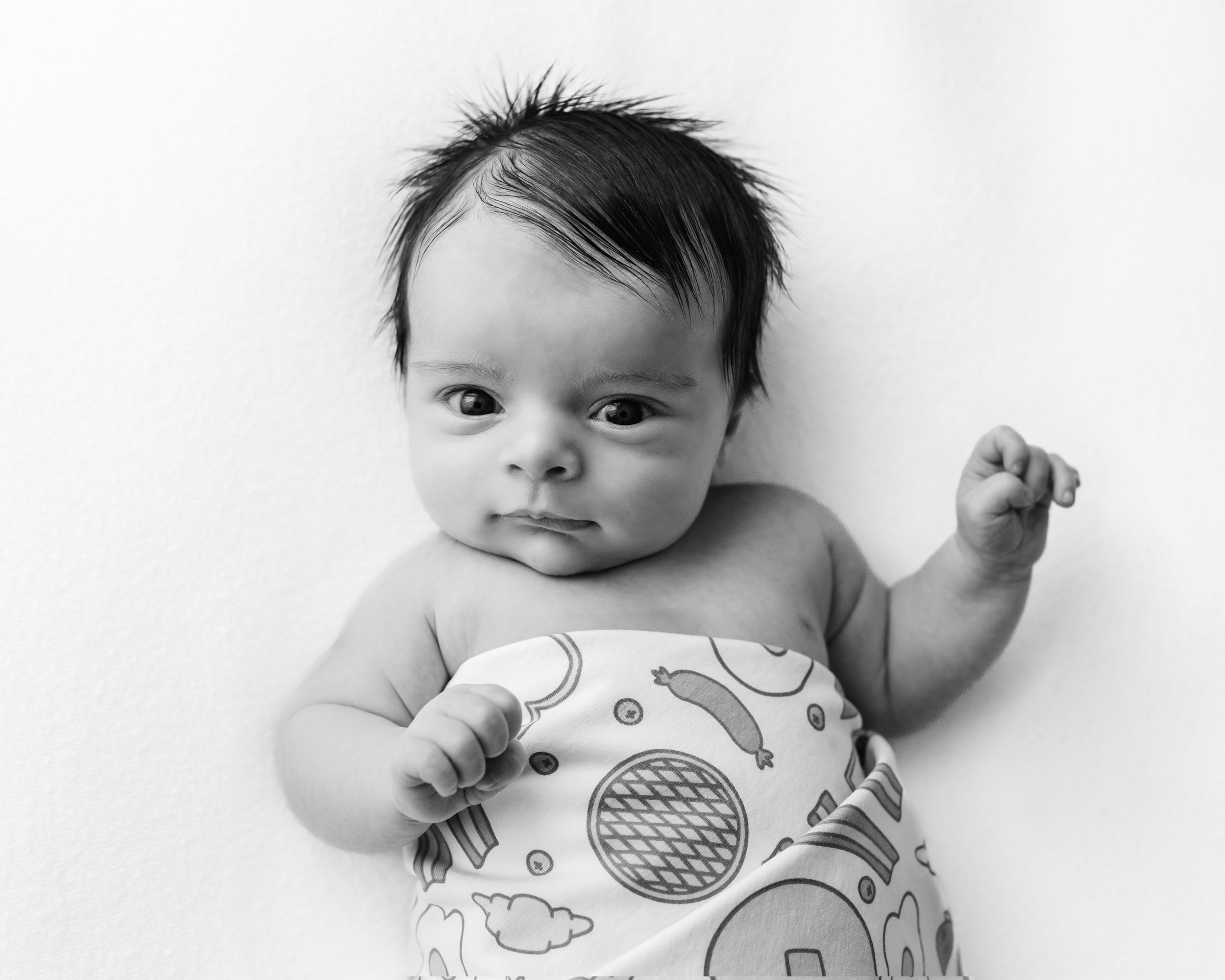 a black and white picture of a baby boy laying on a white backdrop wrapped in a bacon and eggs swaddle blanket looking directly up at the camera during a Pottstown newborn photoshoot