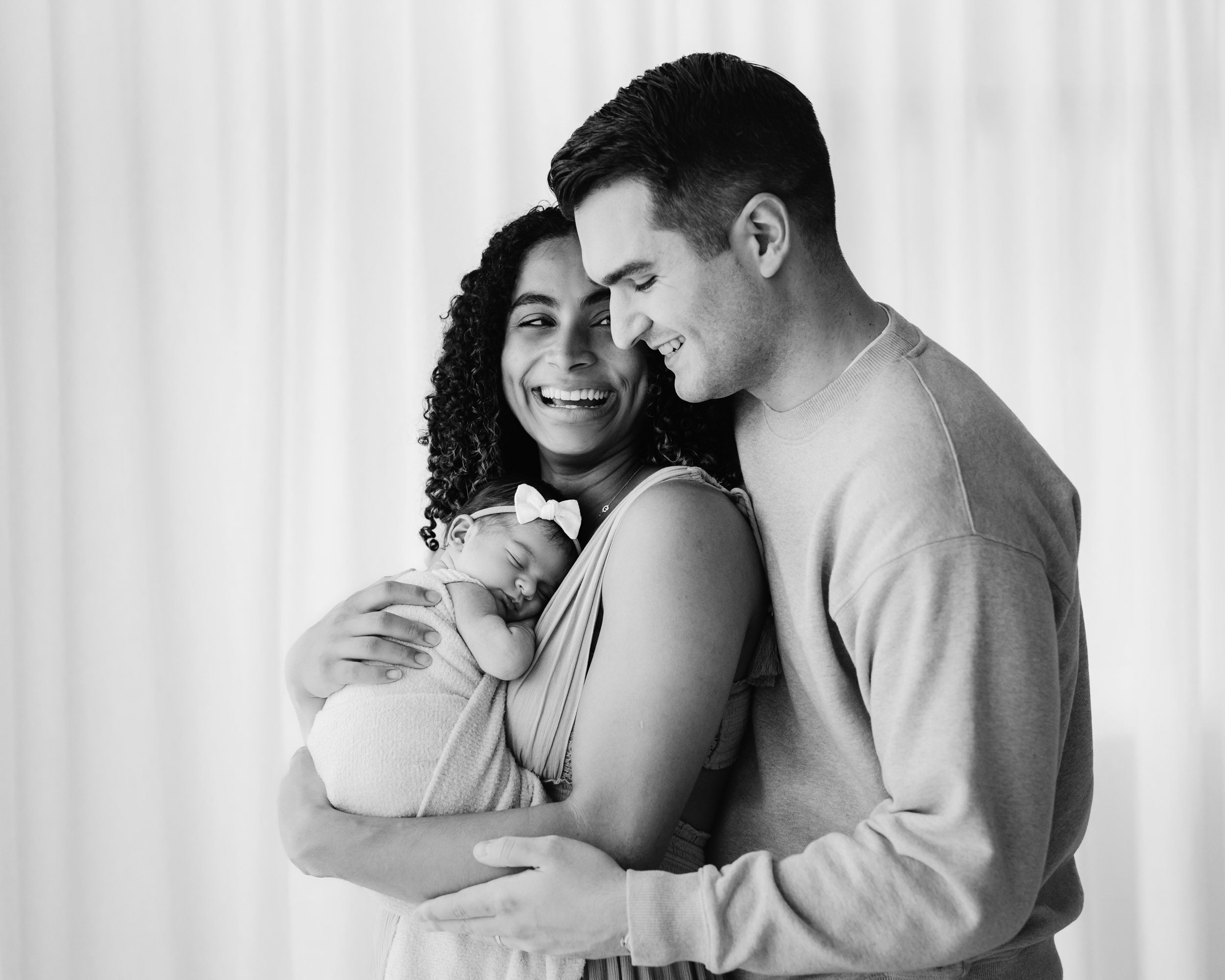 a black and white picture of a mom holding her newborn baby girl against her chest and looking back toward dad who's hugging her from behind during a natural light newborn photo session