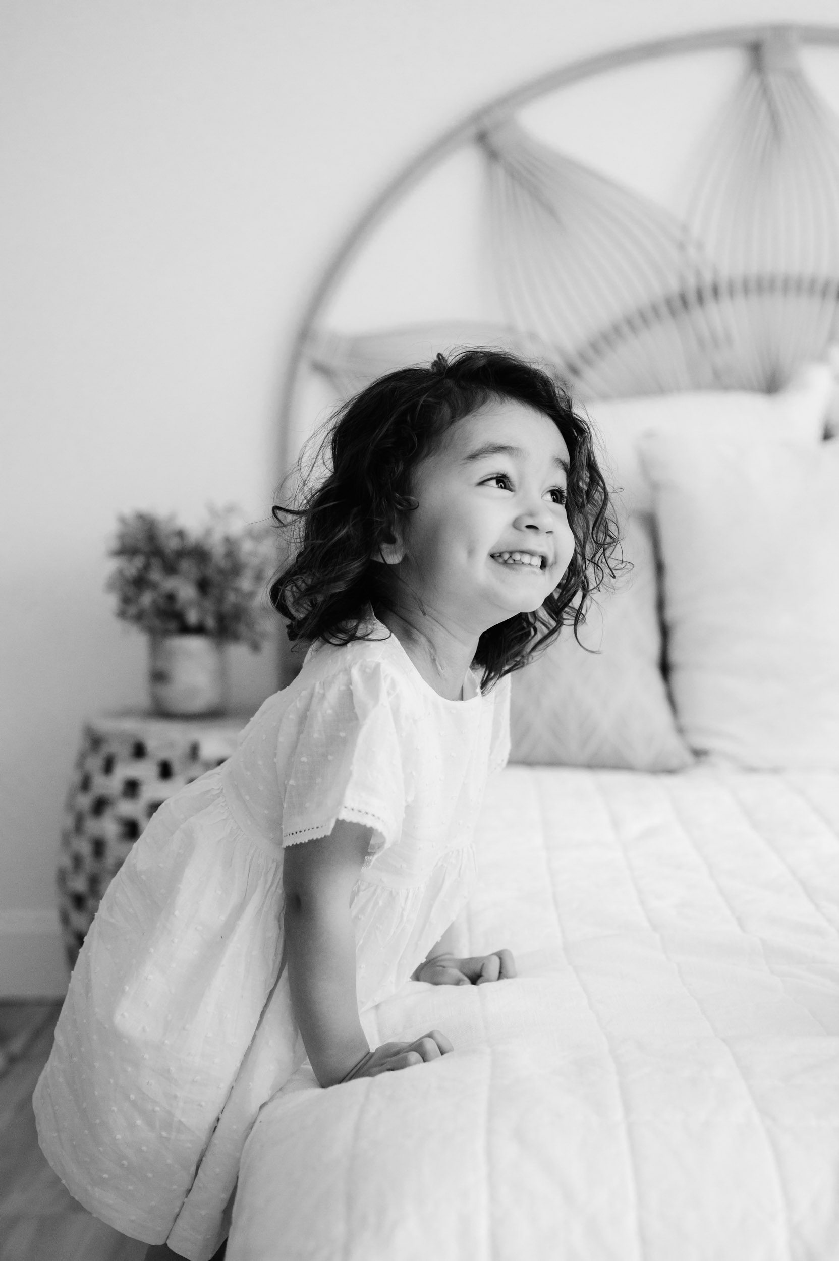 a black and white picture of a young girl leading against the edge of a bed and looking up at her parents with a huge grin on her face during a newborn photo session