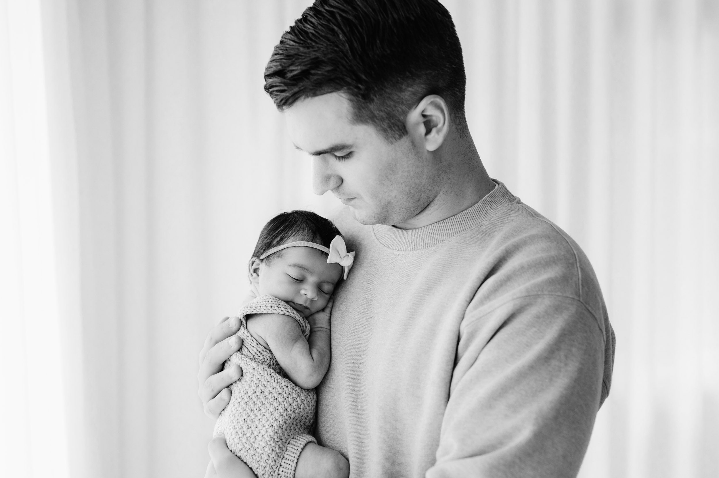 a black and white picture of a new dad holding his baby girl against his chest and looking down at her as she sleeps during a natural light newborn photo session