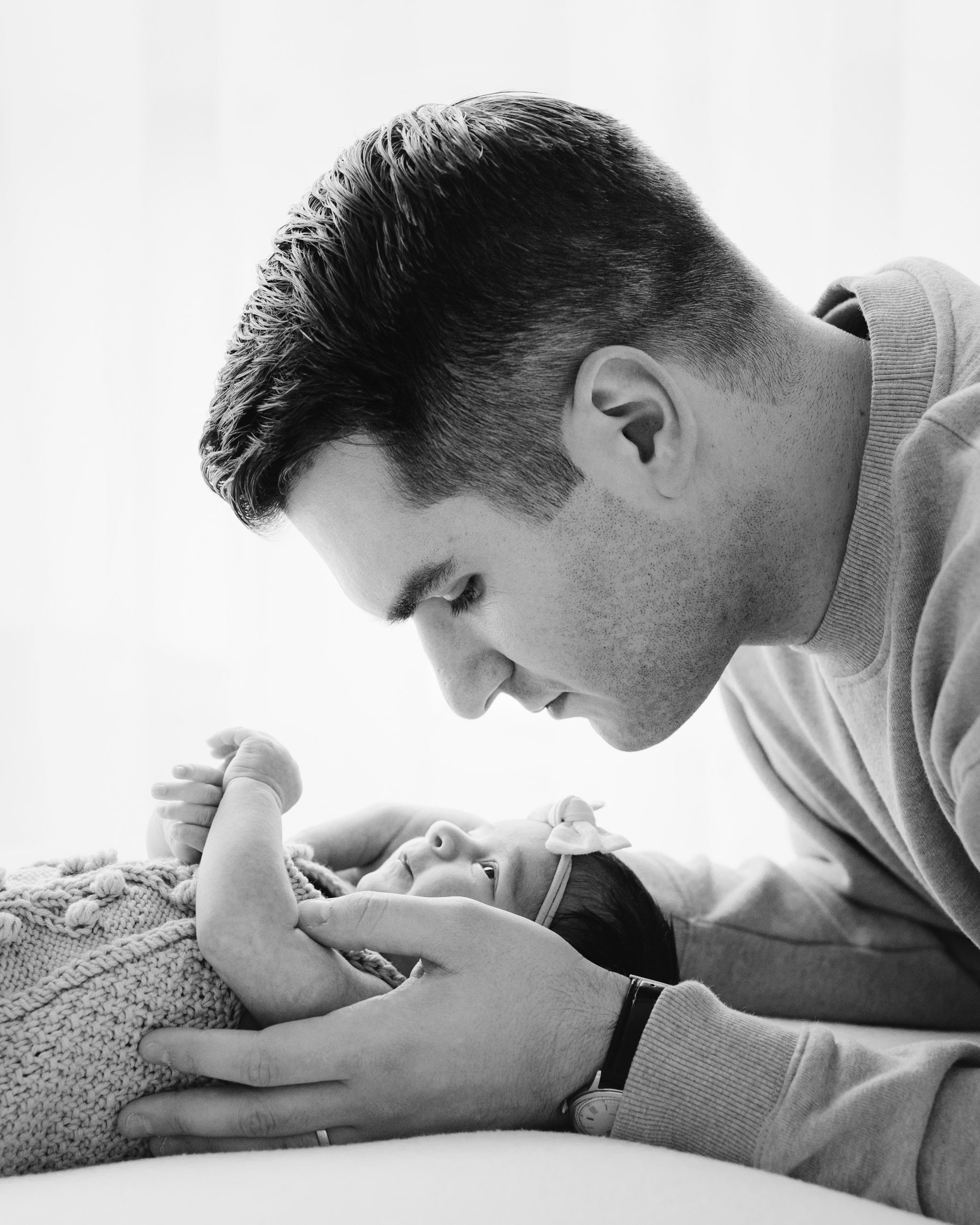 a black and white backlit photo of baby girl laying on a beanbag as her dad cradles her in his hands and gazes down at her from above during a natural light newborn photo session