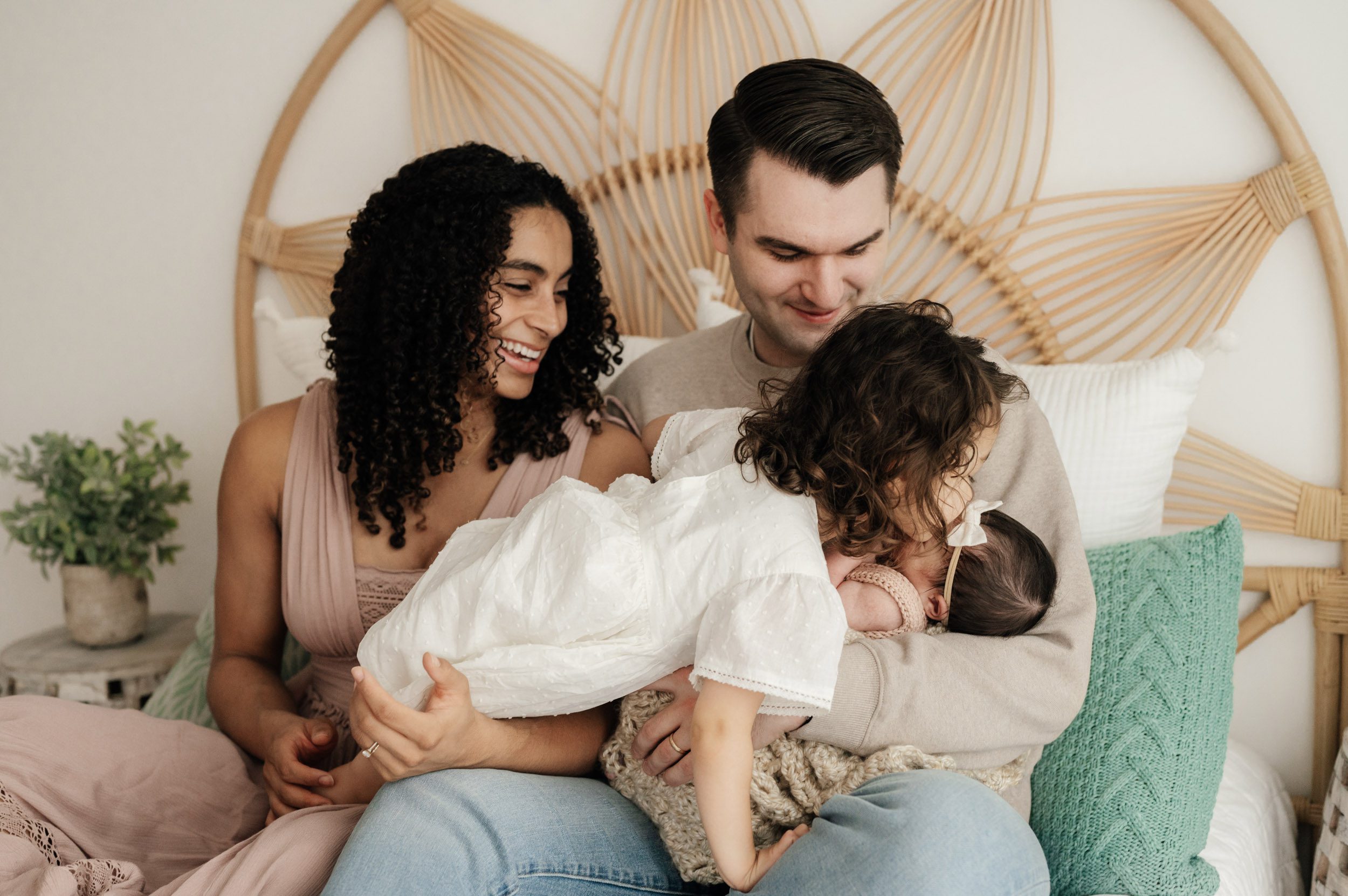 parents sitting on a bed holding their newborn baby girl and older daughter on their lap as big sister climbs across dad's lap and gives her baby sister a kiss on the head during a newborn photoshoot
