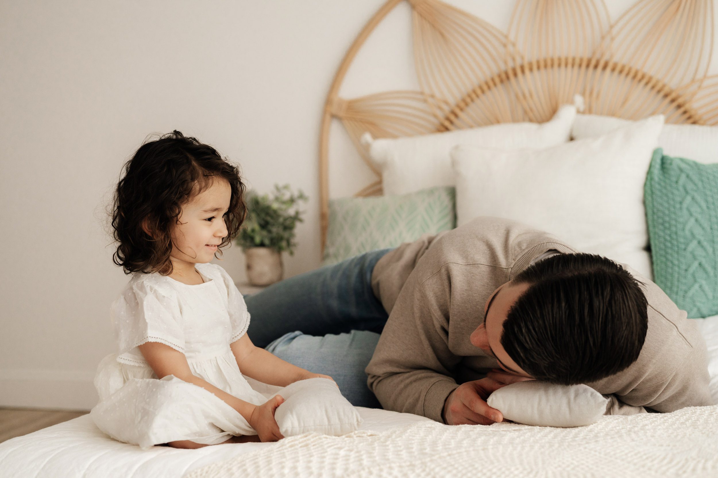 a young girl sitting on a bed and smiling at her dad as he rests his head on a tiny pillow during a newborn photoshoot