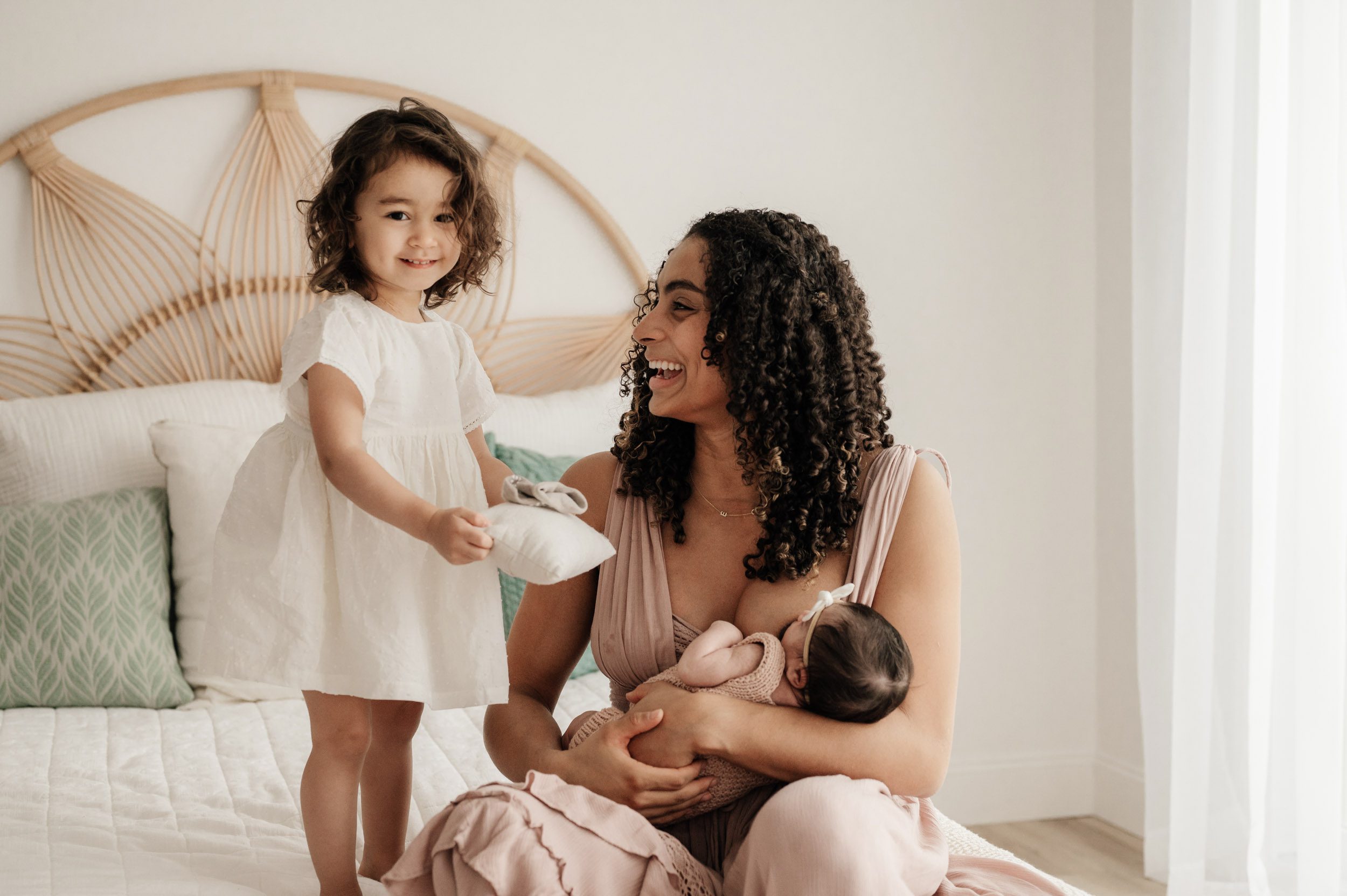 a mom sitting on a bed nursing her baby girl and smiling at her older daughter who is handing her mom a tiny pillow for the baby during a newborn photo session