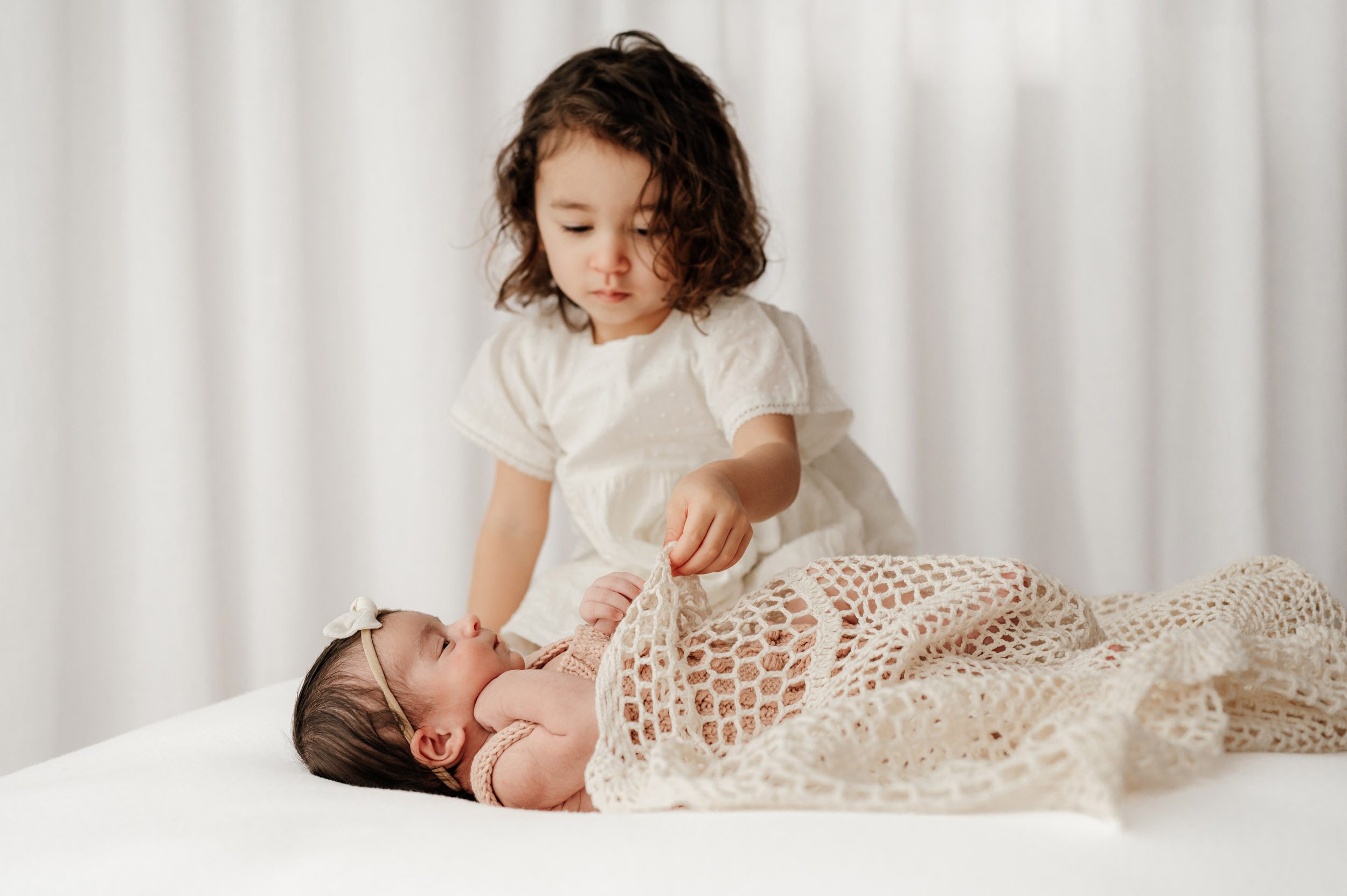 a newborn baby girl laying on a white bean bag while her older sister gently covers her in a crochet knit blanket during a newborn photo session