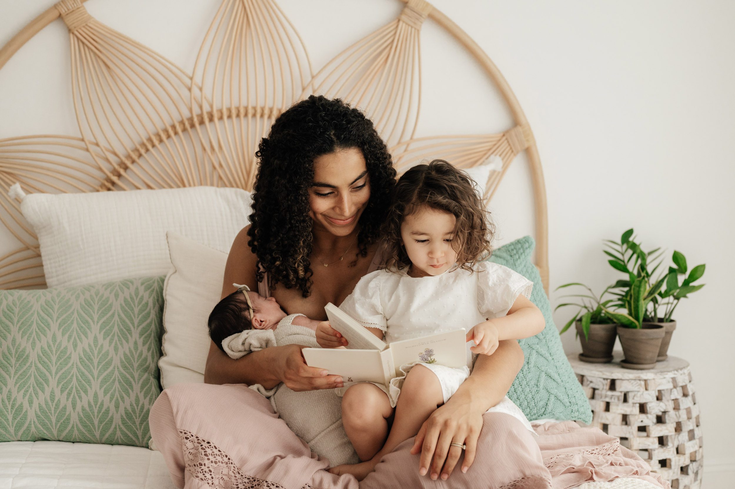 a mom sitting on a bed and nursing her newborn baby girl while also holding her older daughter on her lap and reading a book to her during a natural light newborn photo session